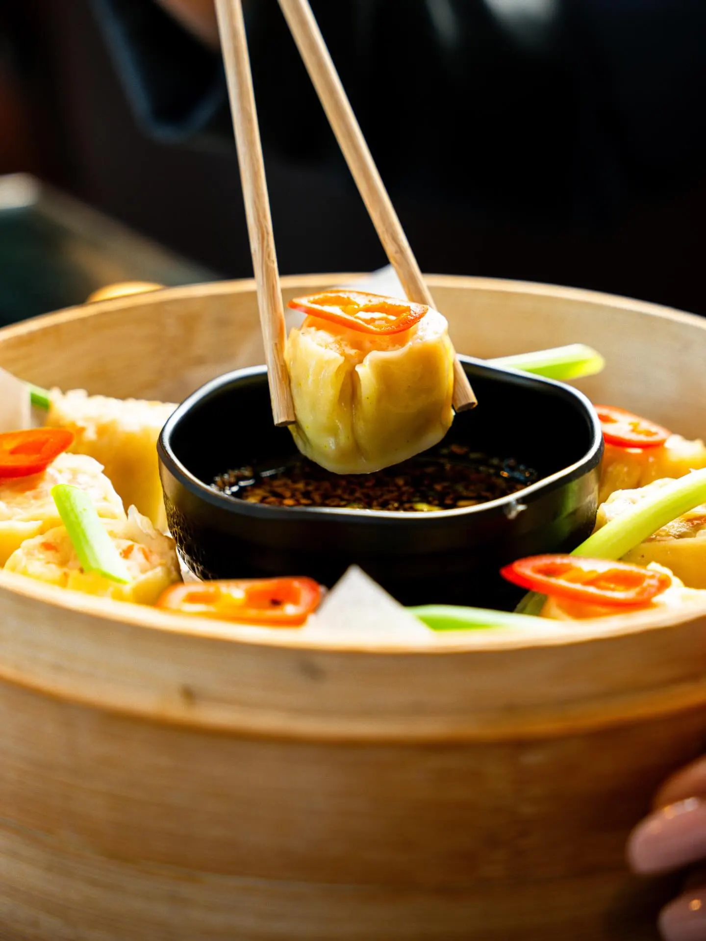 A dumpling lifted on chopsticks above a small pan of broth and vegetables