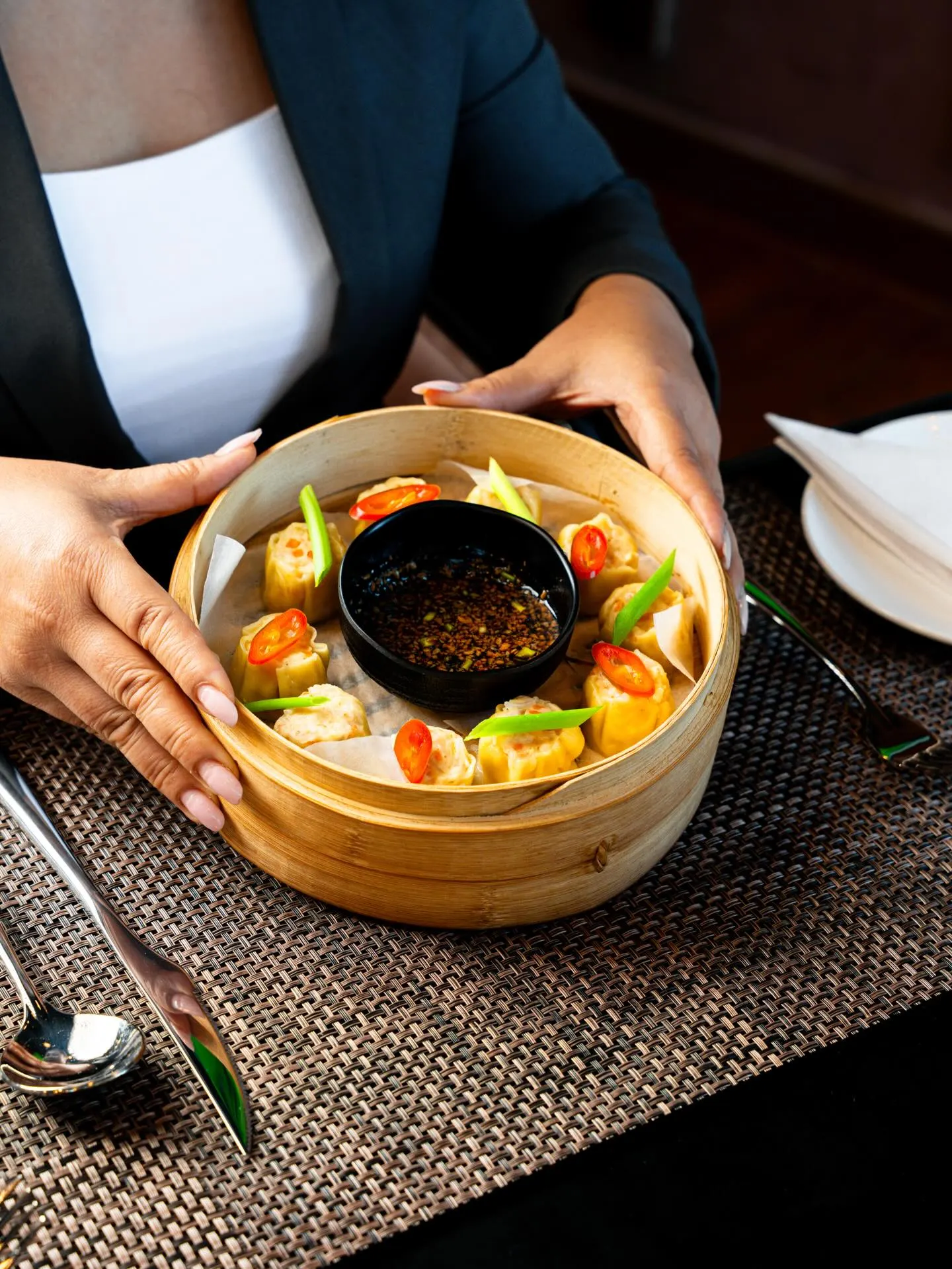 A bamboo steamer of vegetables and broth on a dark table, hands resting either side