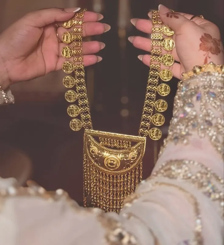 A bride's hennaed hands holding up a traditional gold coin necklace above a beaded, embroidered sleeve