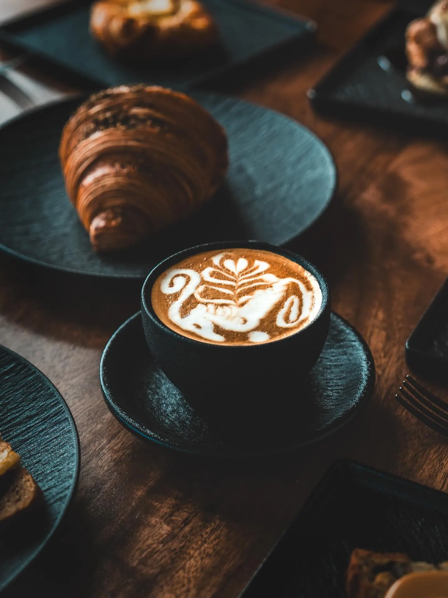 A croissant on a dark plate beside a coffee with poured latte art, on a dark table