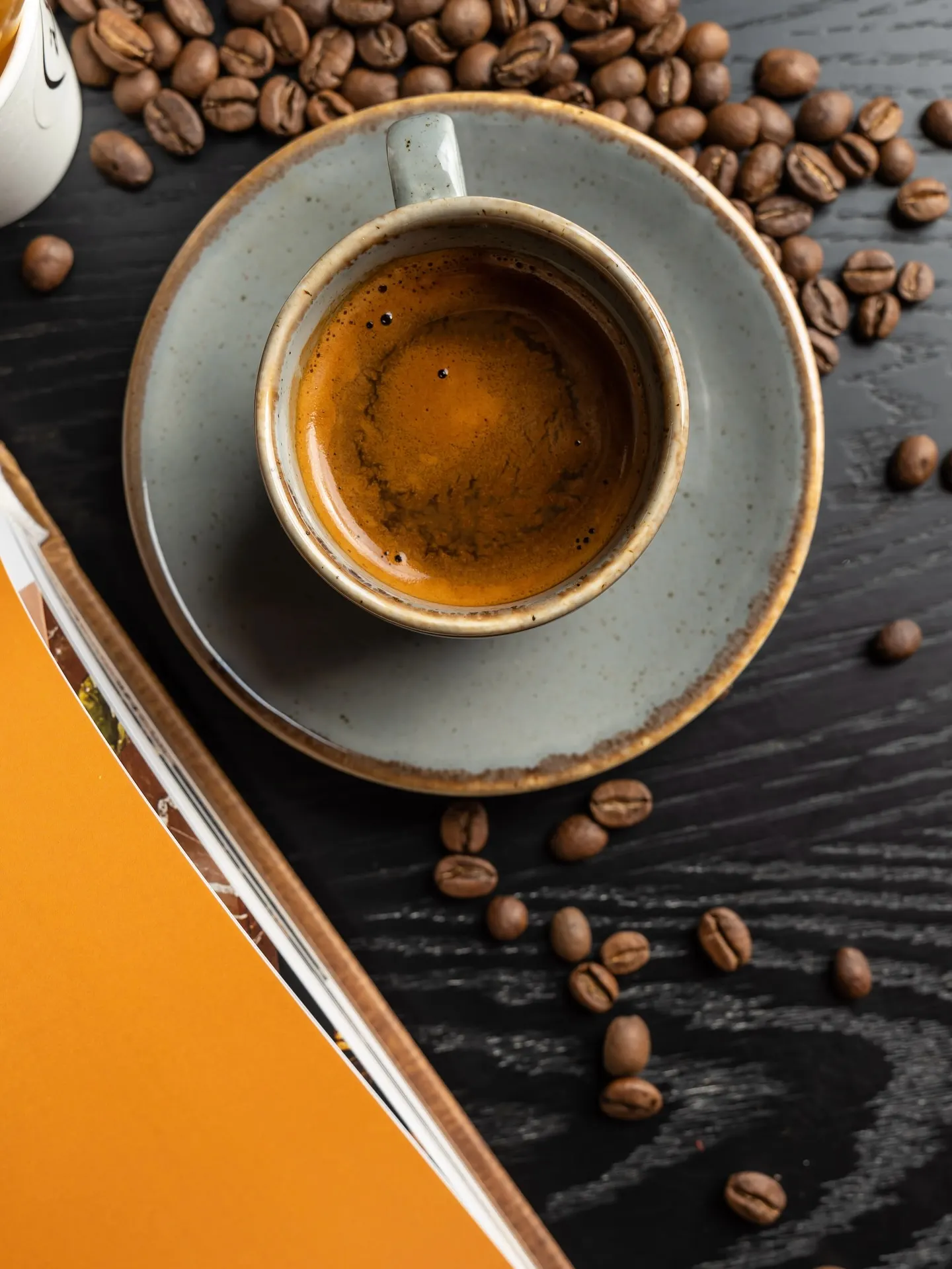An espresso in a grey cup on a bright orange surface, coffee beans scattered beside it