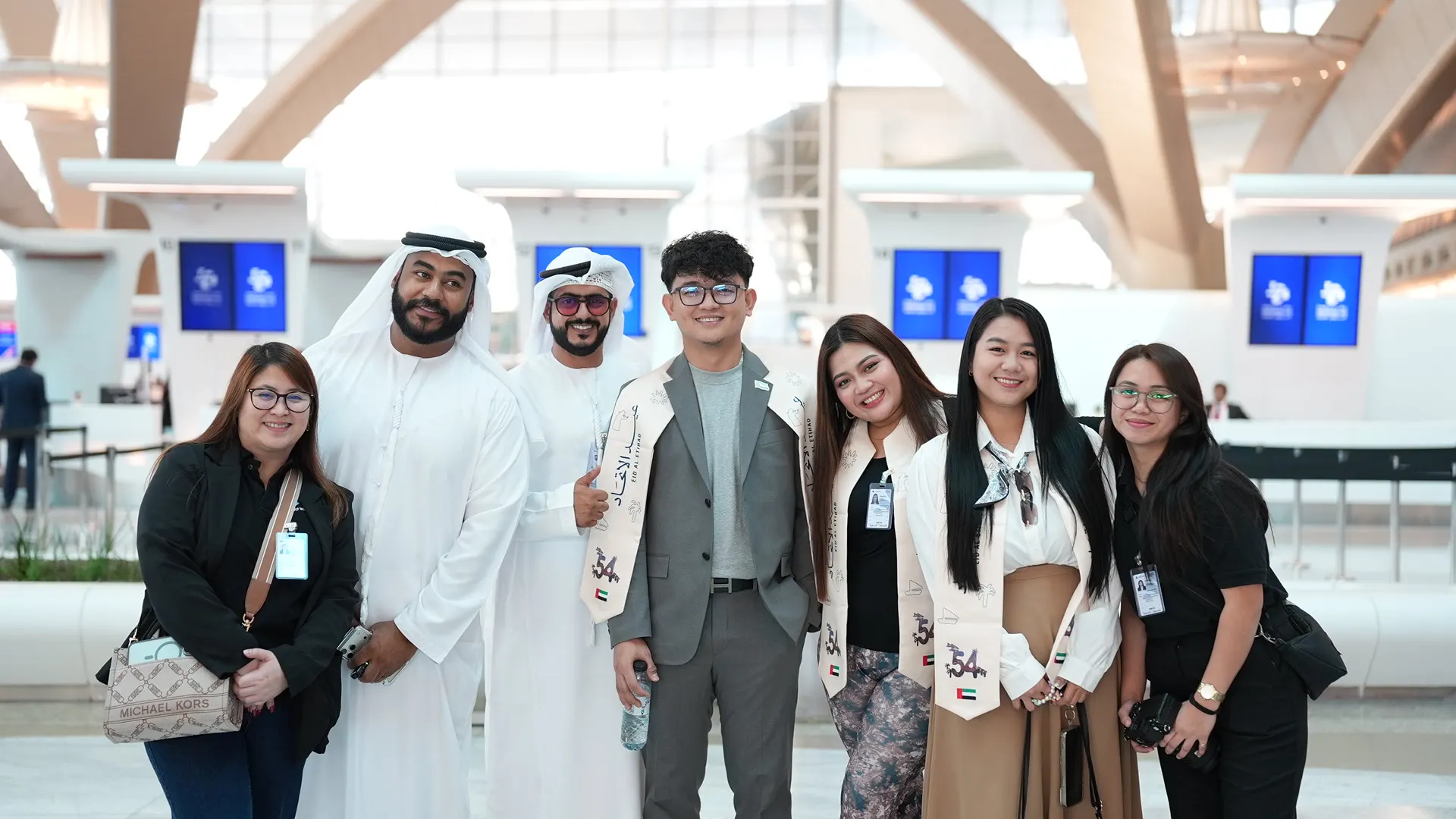 Eight people posing together in an airport terminal, several wearing Eid Al Etihad sashes