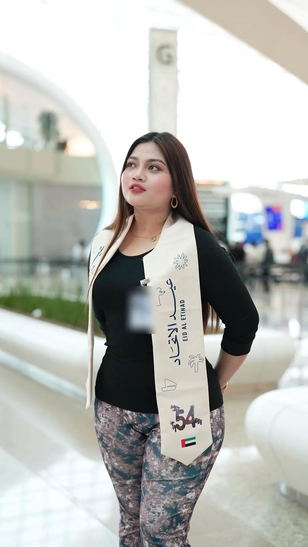 A woman standing in an airport terminal wearing an Eid Al Etihad sash