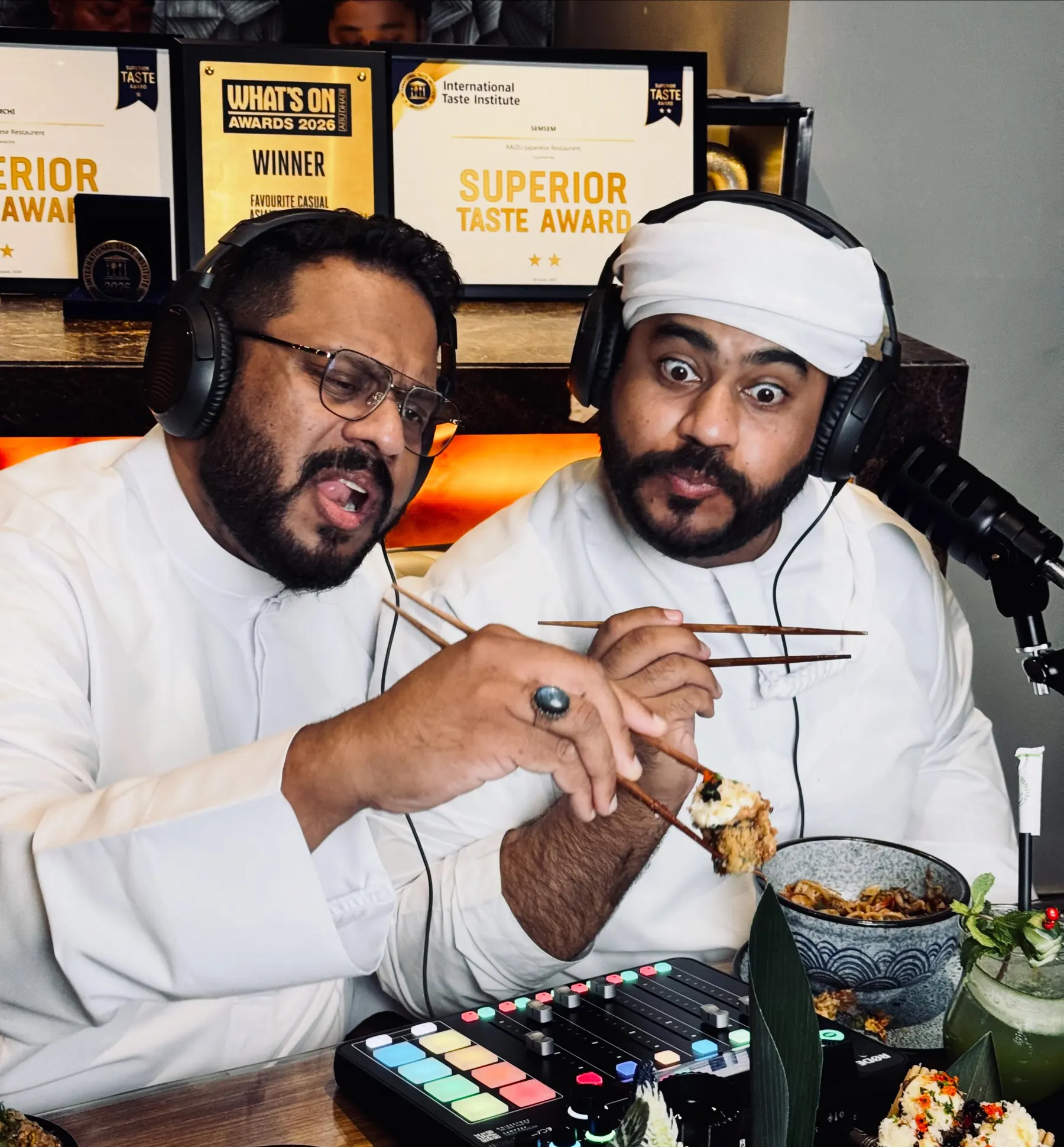 Two podcast hosts eating at a restaurant table with microphones