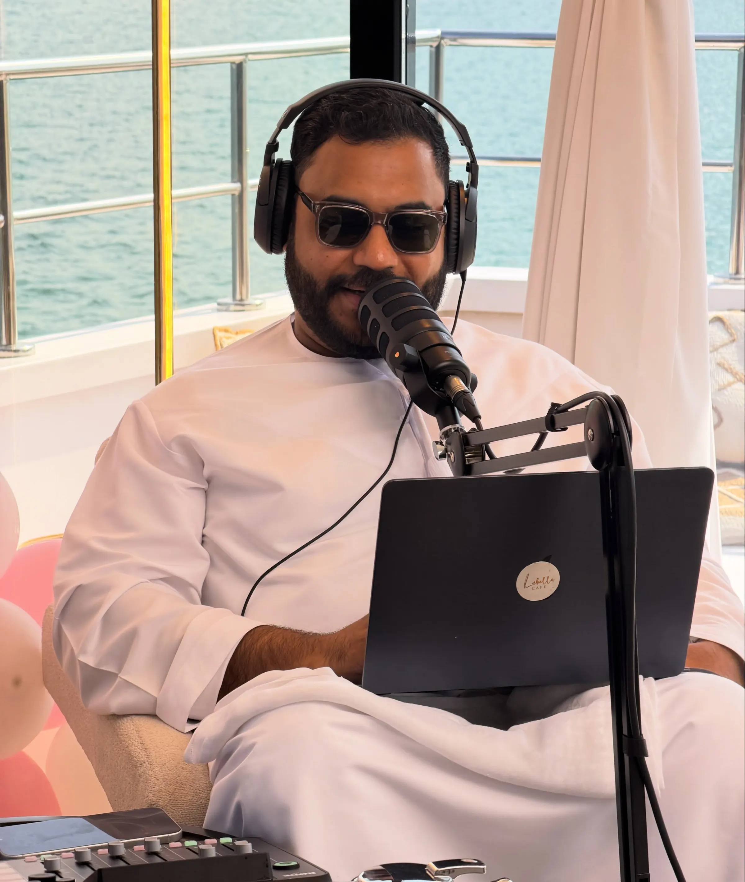 Podcast host in a kandura and headphones at a microphone with a laptop