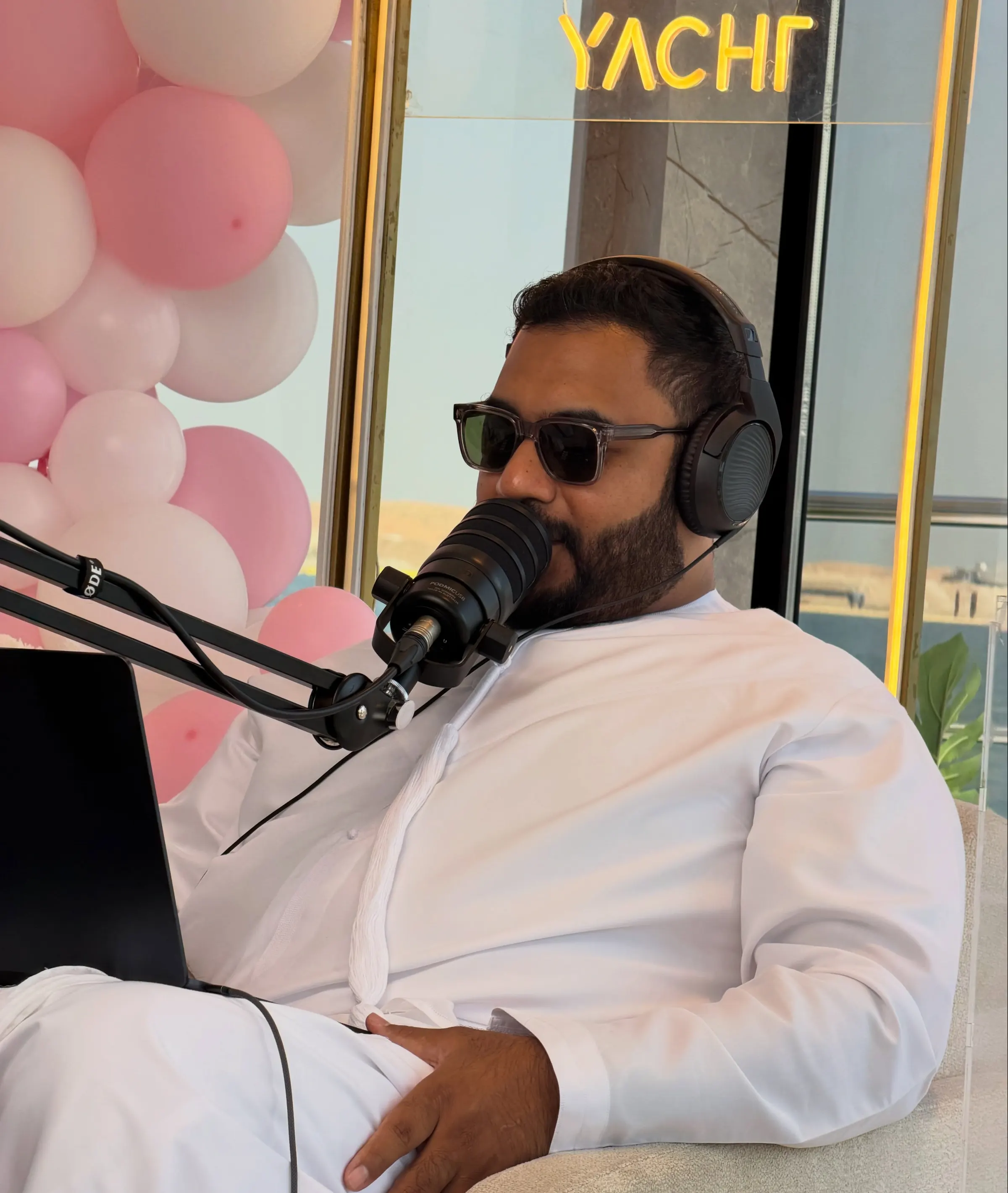 Podcast host at a microphone on a yacht, pale balloons behind