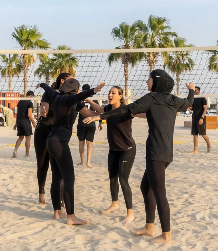 Volleyball players in black kit high-fiving one another on the sand in front of the net, with palm trees behind them