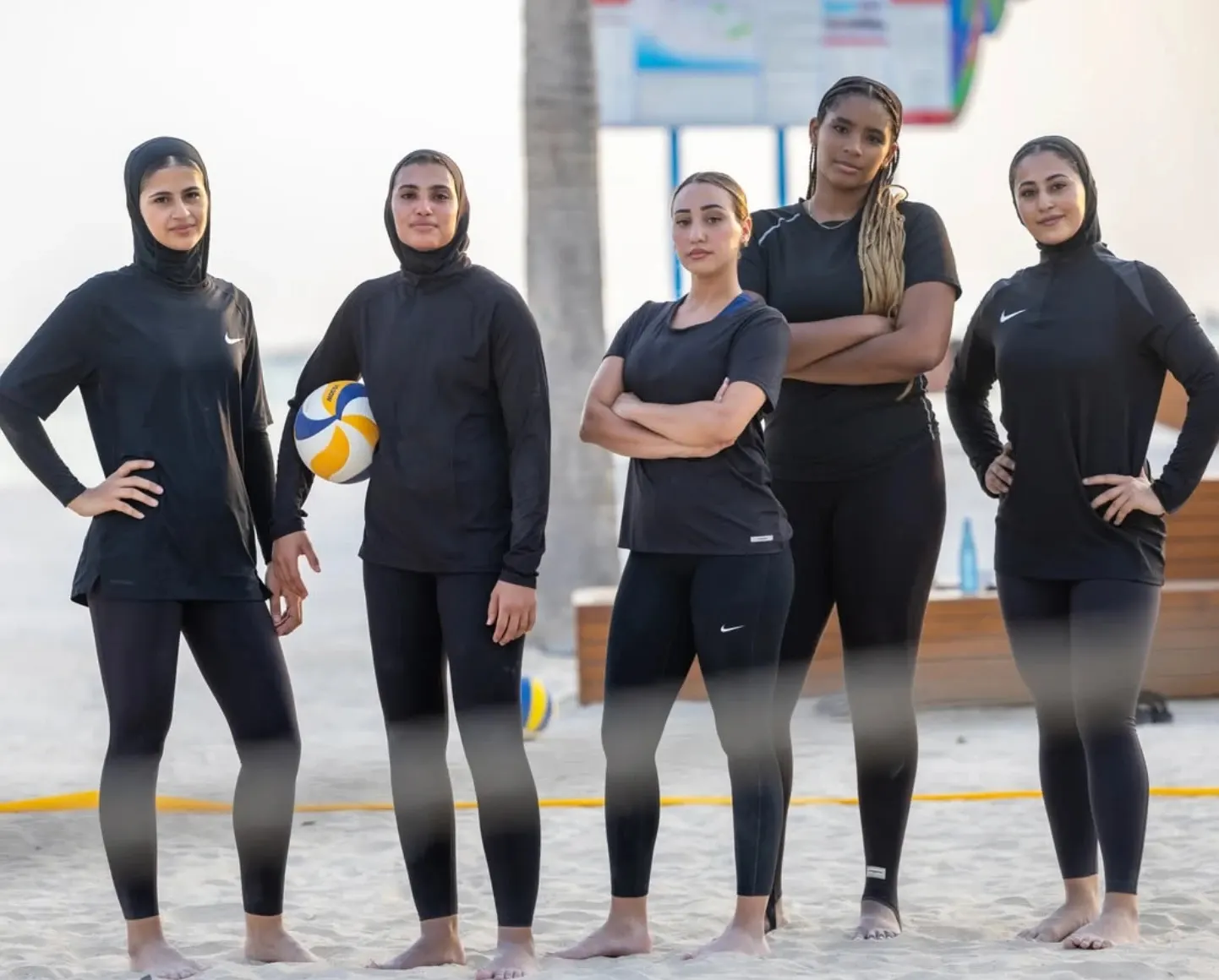 Five women's volleyball players in black training kit standing on the sand of a beach court, one holding a volleyball, with a palm tree behind them