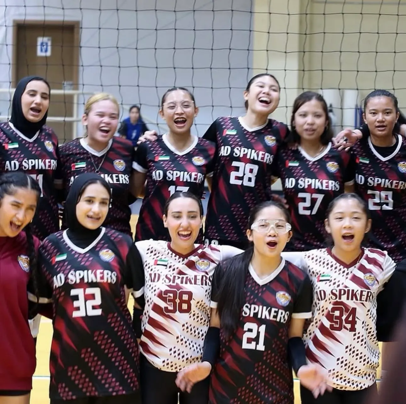 AD Spikers players in maroon, black and white team jerseys cheering together, arm in arm, in front of a volleyball net