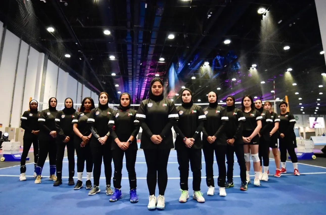 The AD Spikers squad in black kit lined up in two rows on a blue indoor court under the lights of a large exhibition hall