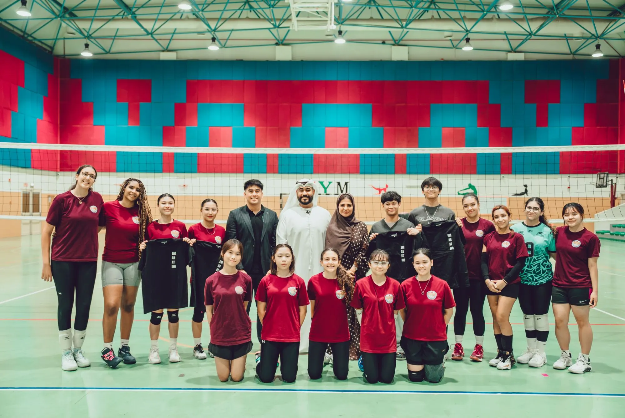 The AD Spikers squad in maroon and red team shirts lined up in front of a volleyball net with Fahad Al Blooshi, in a white kandura, and the Evo Media Pro team, several of them holding up black Emaratino T-shirts