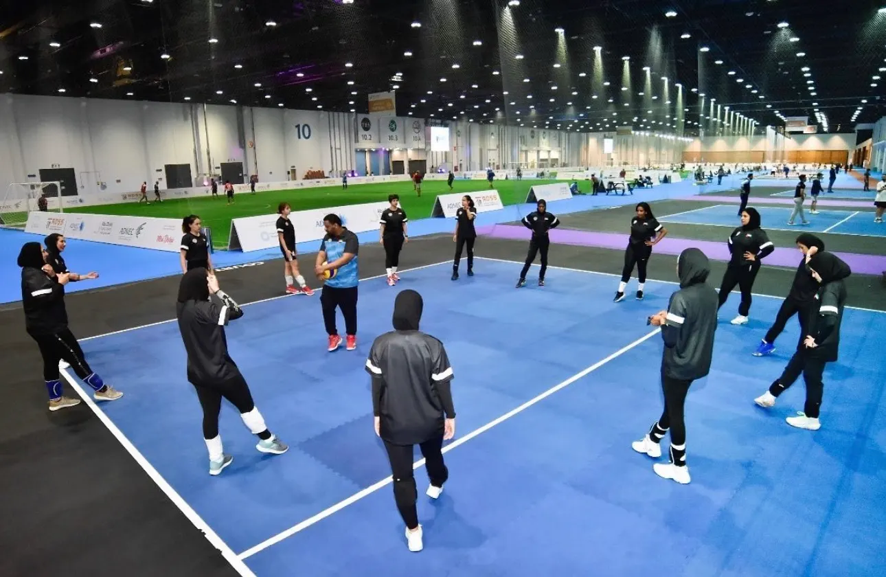 AD Spikers players in black kit standing in a circle on a blue indoor court around a man in a blue top holding a volleyball, with more courts behind them in a large hall
