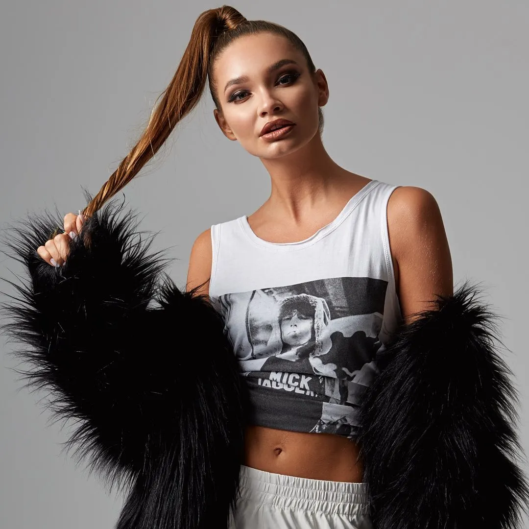 A fashion portrait of a model in a black fur coat and printed vest against a pale studio wall