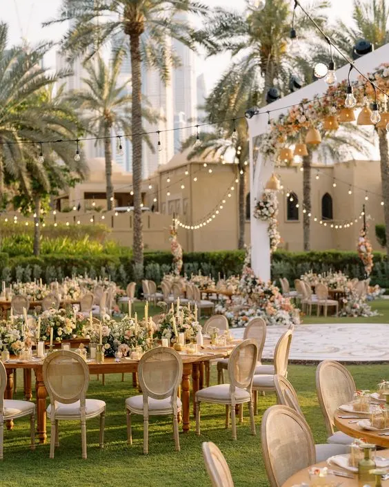 An outdoor wedding reception set for dinner on a lawn under palm trees and string lights, with floral arches, wooden tables and cane chairs