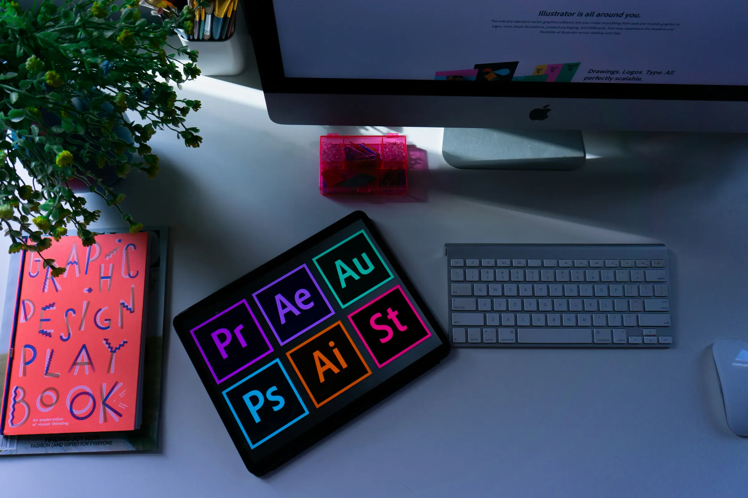 Overhead view of a design desk: a tablet showing creative software icons beside a keyboard, a desktop screen and a graphic design book