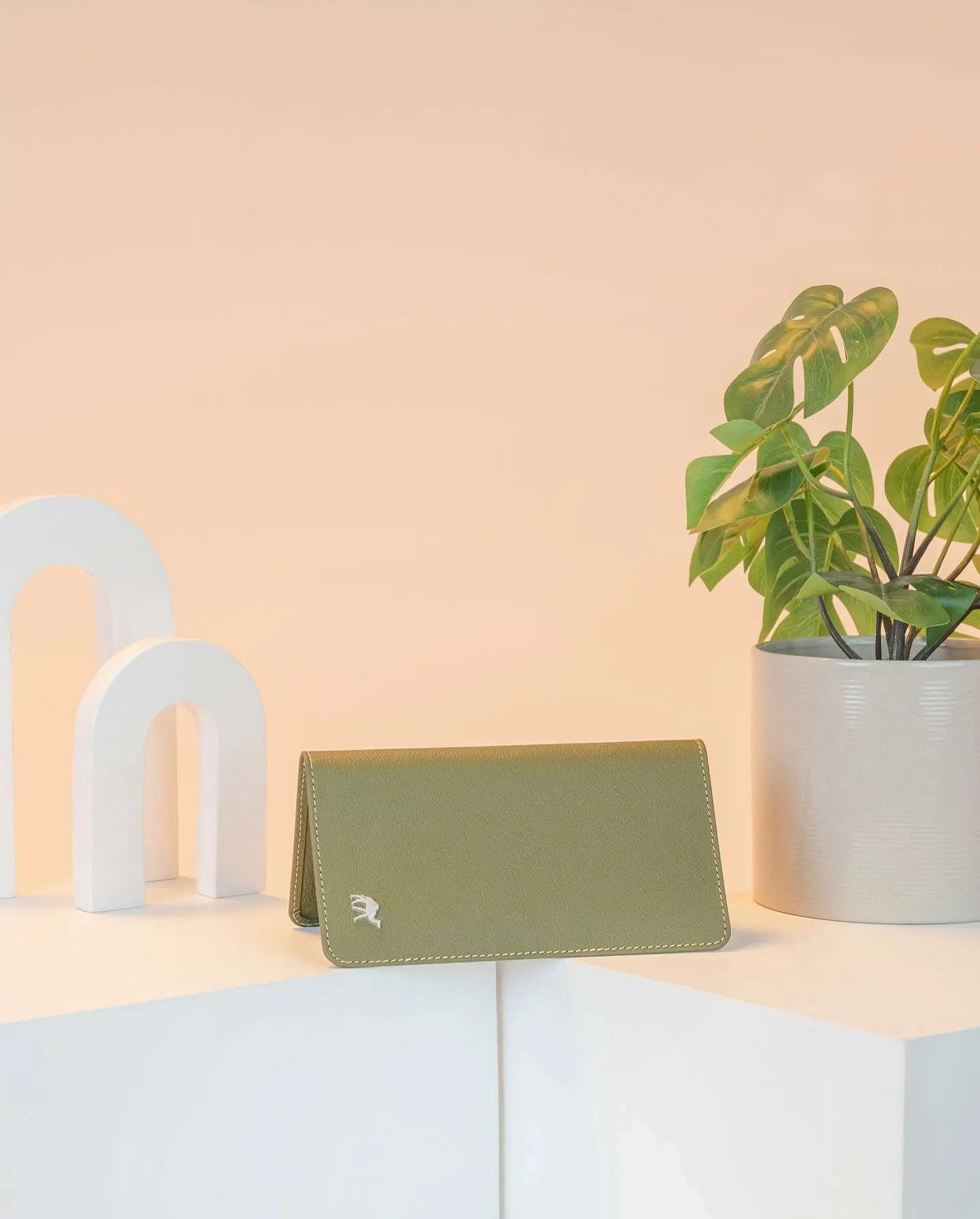 A green leather wallet and its box arranged on pale plinths with potted plants behind