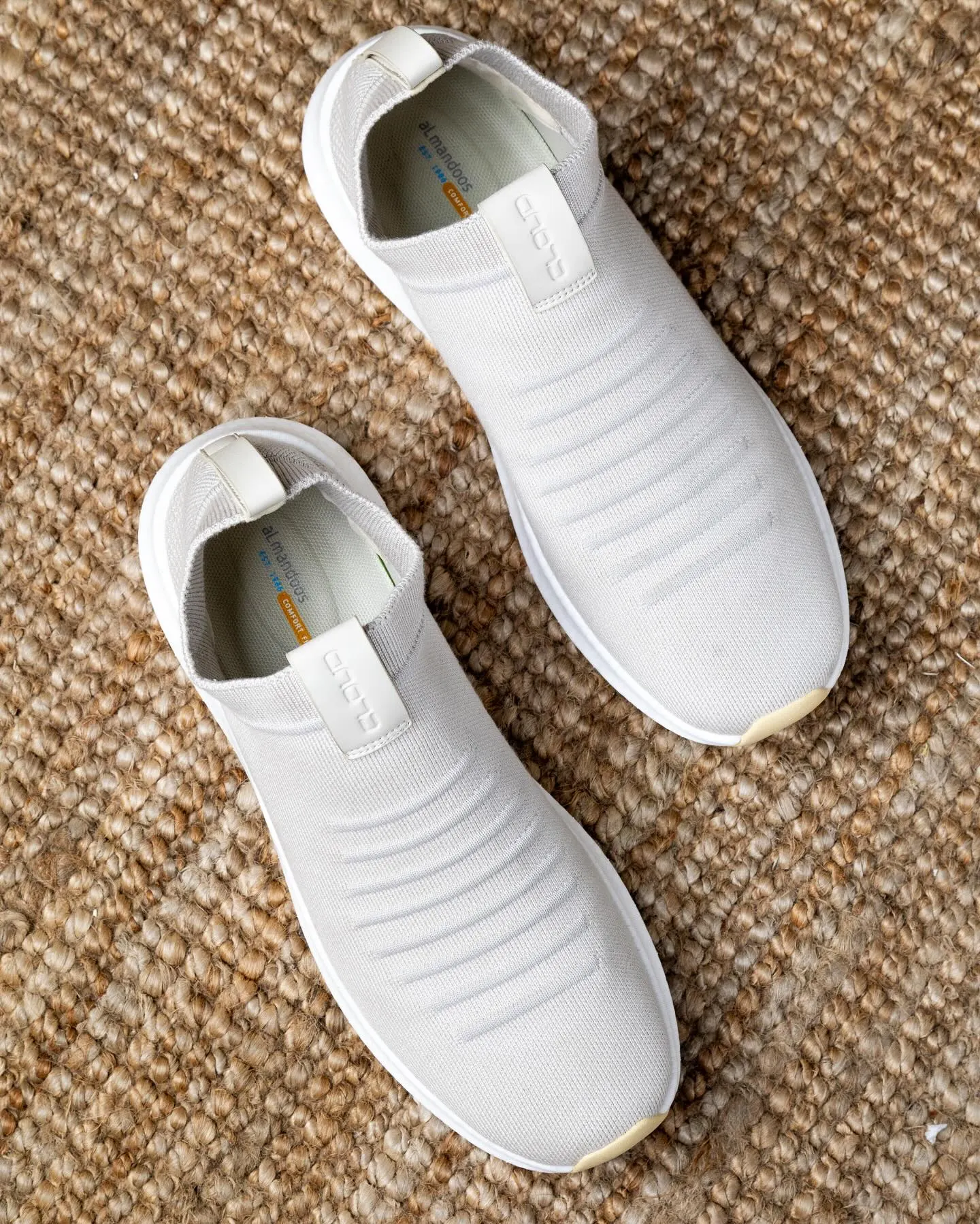 A pair of white knitted slip-on shoes photographed from above on woven matting
