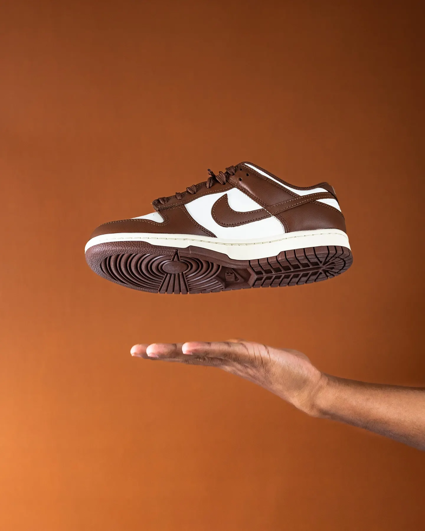 A white and brown sneaker suspended above an open hand against an orange background