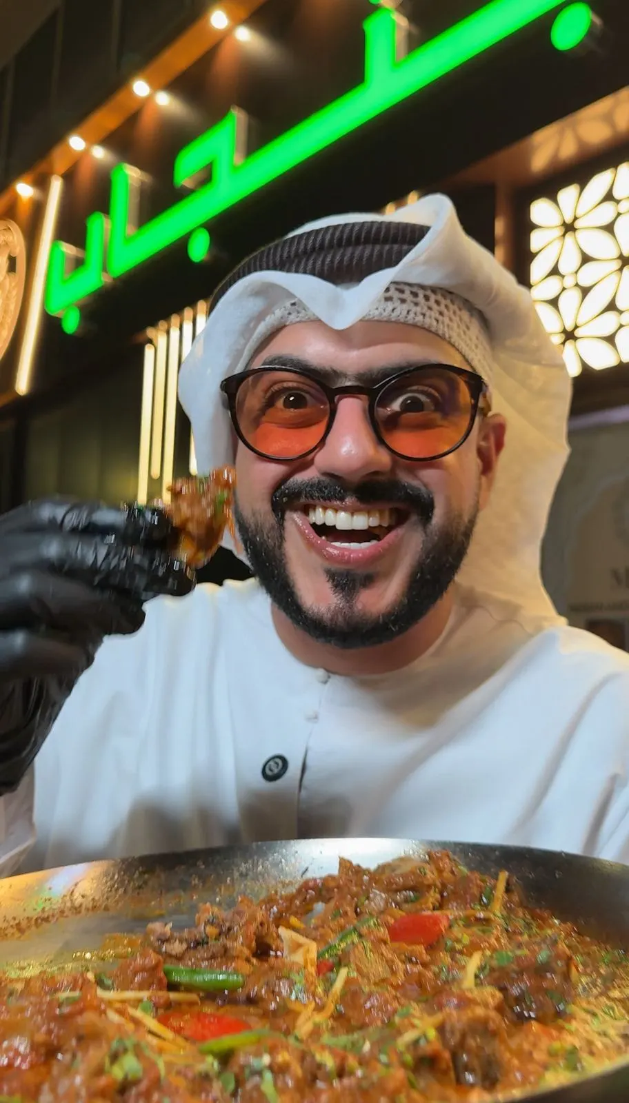 Still from a vertical food video: a presenter in a white ghutra and orange-tinted glasses holds up a bite from a large pan of curry outside a restaurant with a green neon sign