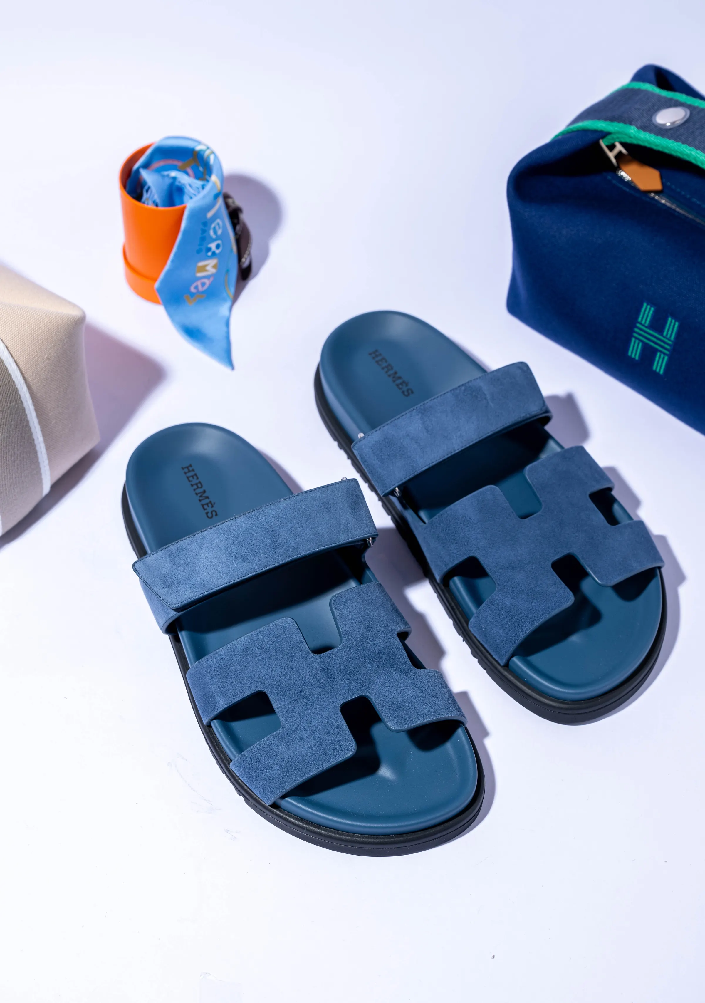 Luxury fashion flat lay: a pair of blue suede sandals beside a silk scarf in an orange box and two canvas pouches, on a white set