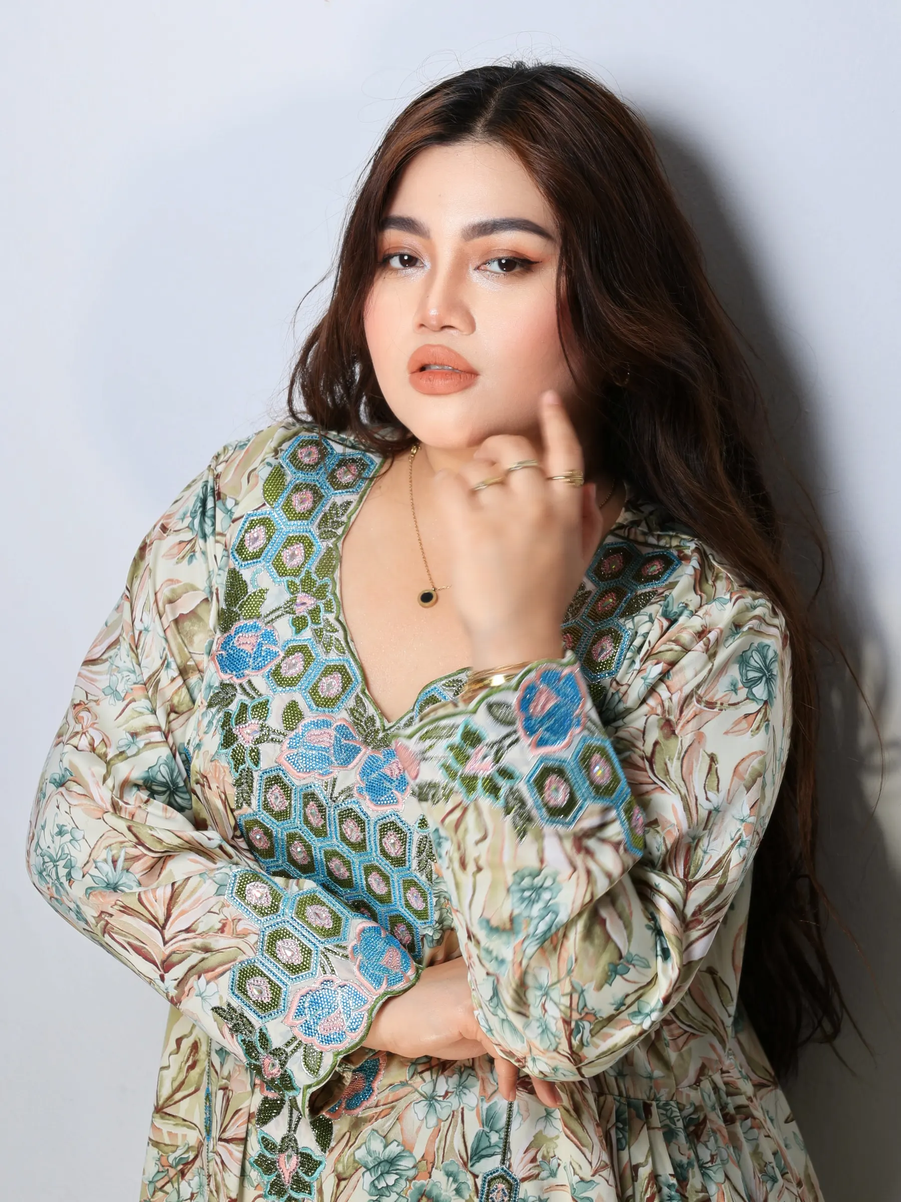 A studio portrait of a woman in a patterned floral dress, hand raised to her chin, against a pale backdrop