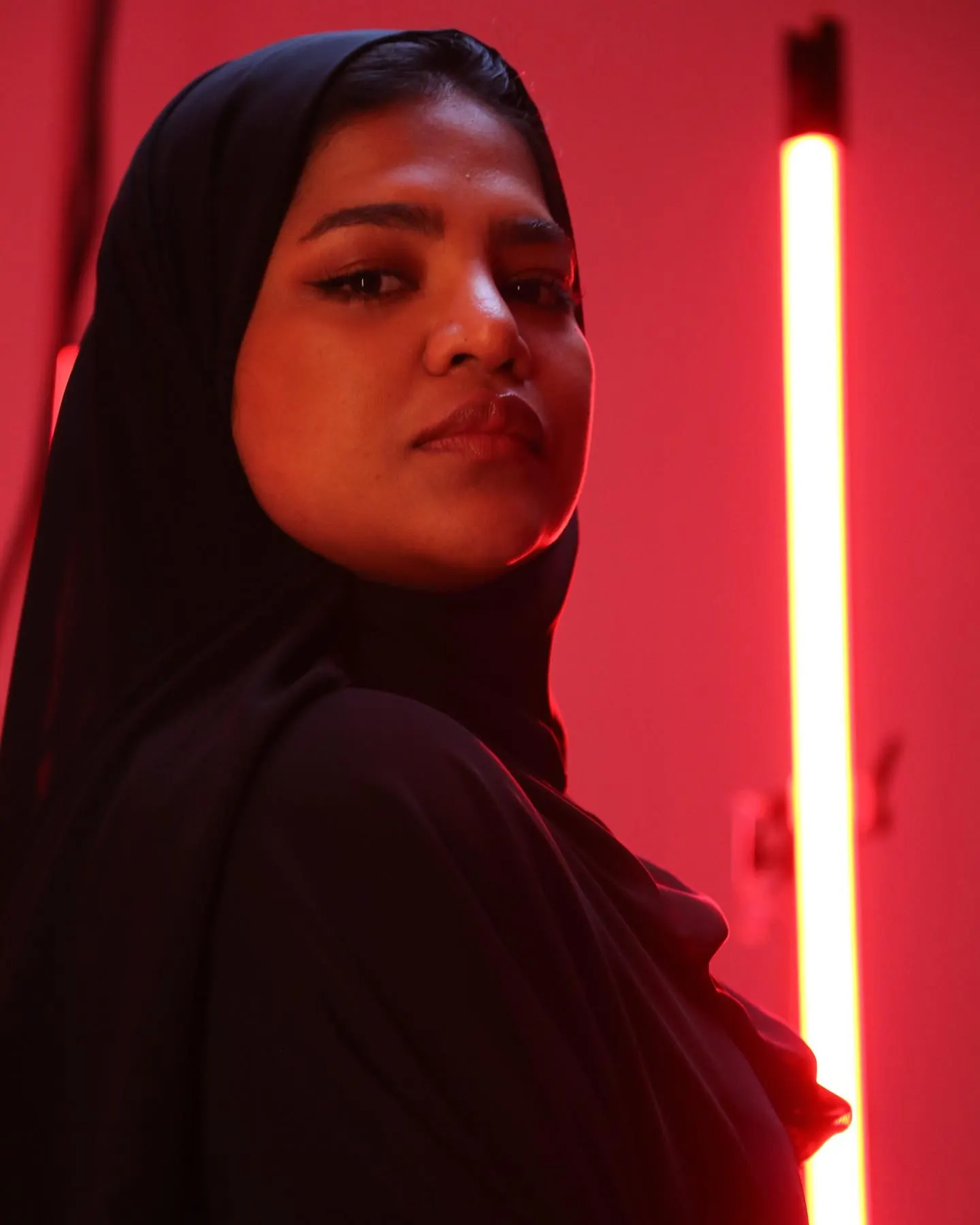 A three-quarter studio portrait under magenta light of a woman in a black hijab