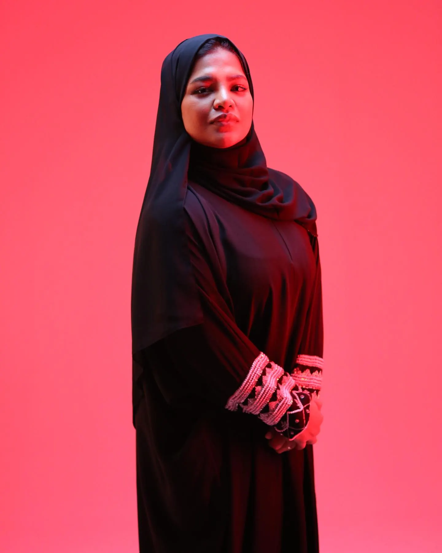 A full-length studio portrait of a woman in black against a magenta backdrop, hands together