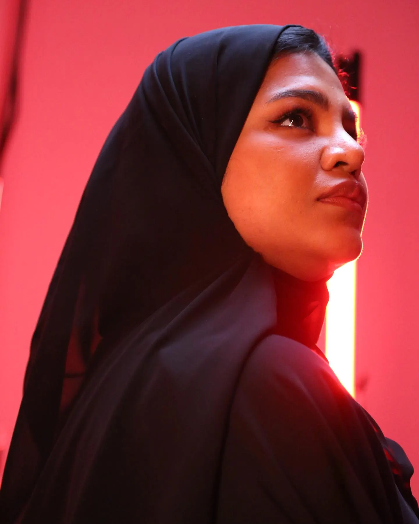 A studio portrait under magenta light of a woman in a black hijab looking upward