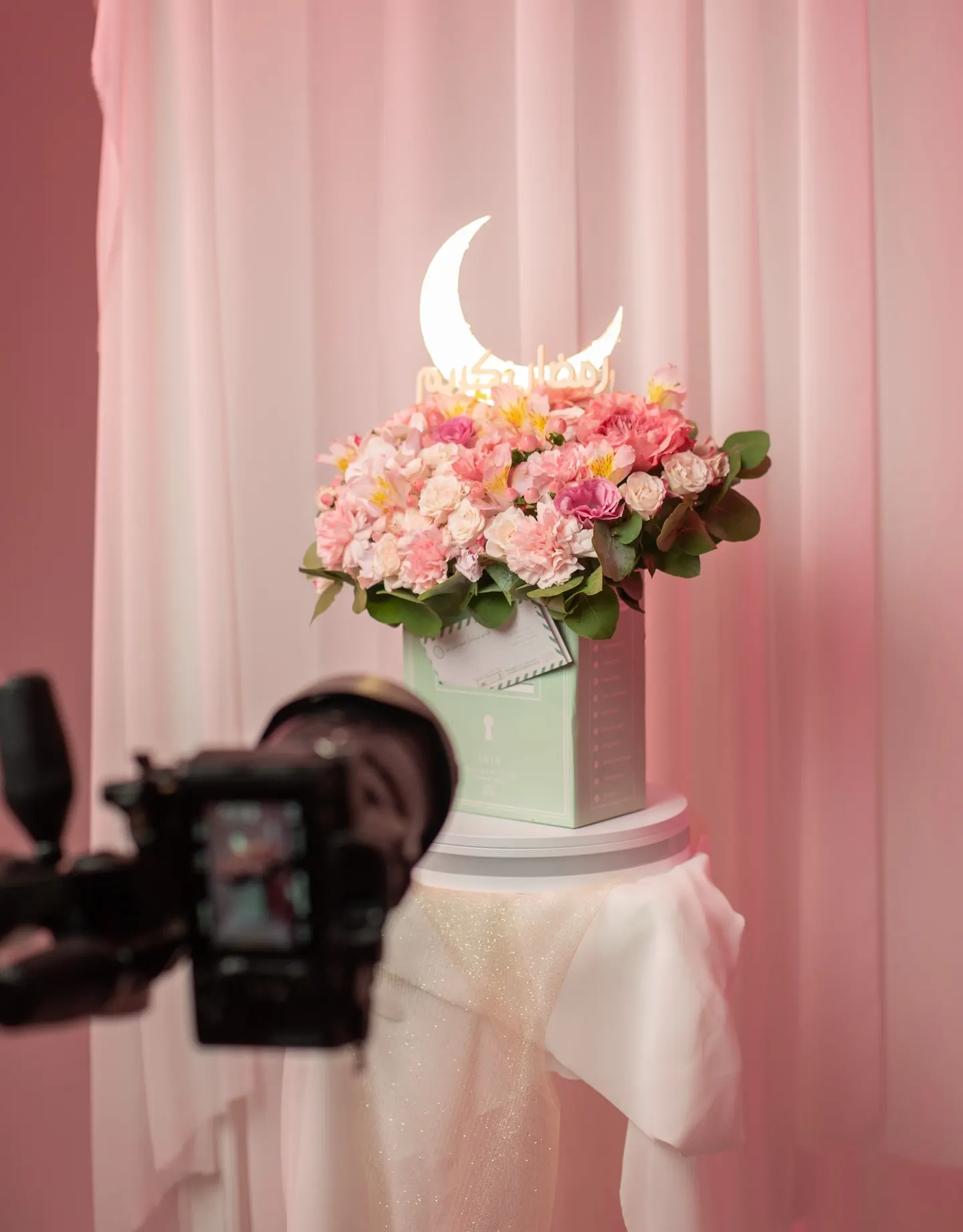 A camera on a tripod framing a bowl of pink flowers on a draped plinth, monitor showing the shot