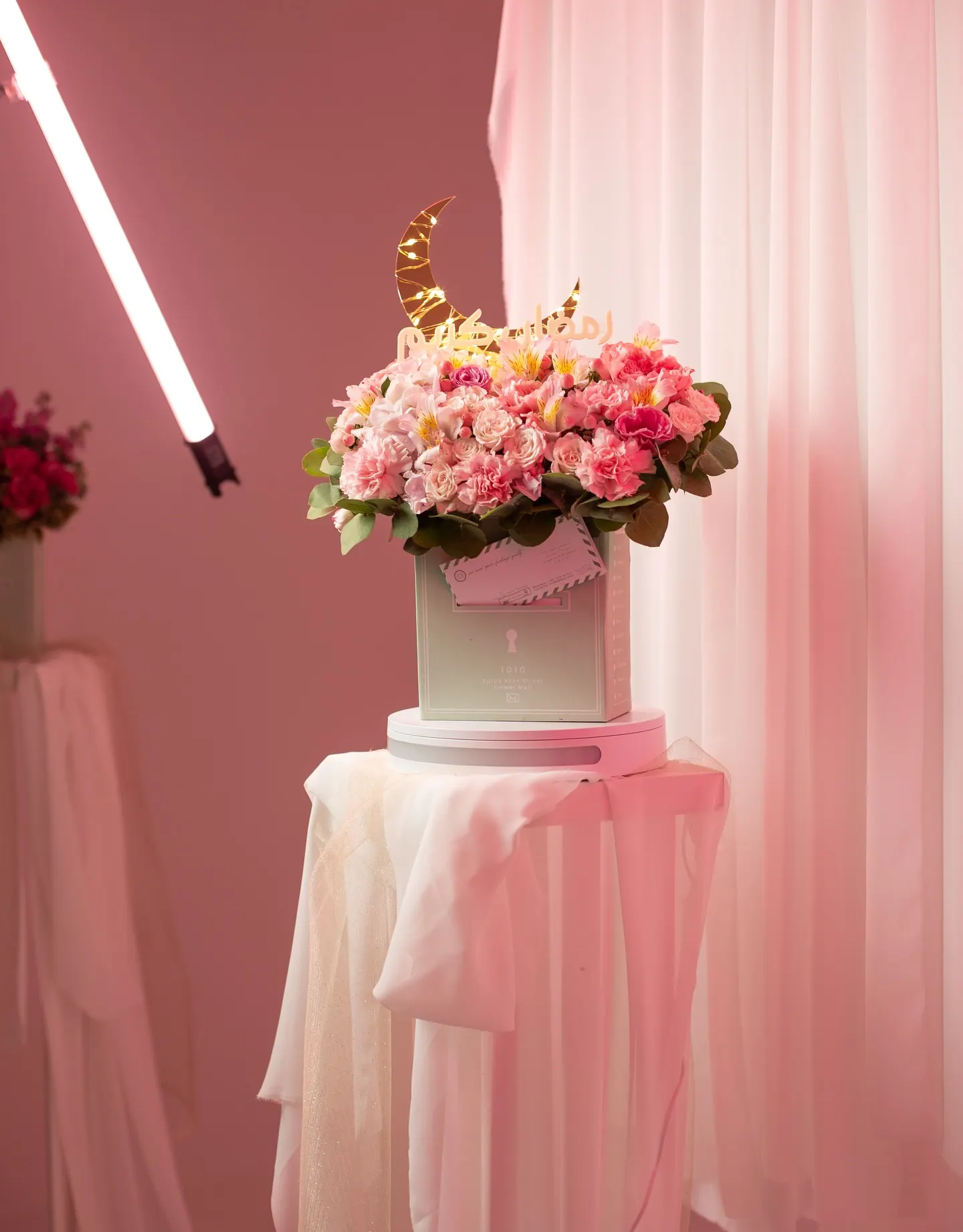 A vertical tube light beside a bowl of pink flowers on a draped plinth, softbox overhead