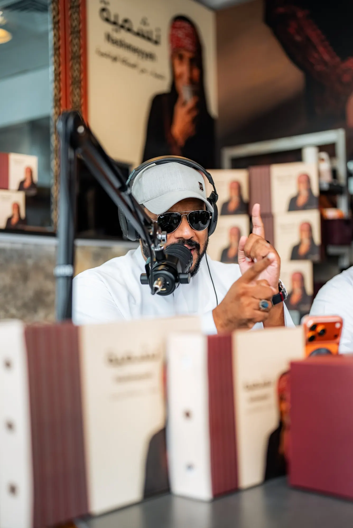 A podcast host in a grey cap, aviator sunglasses and headphones making a point at the microphone, product boxes blurred in the foreground
