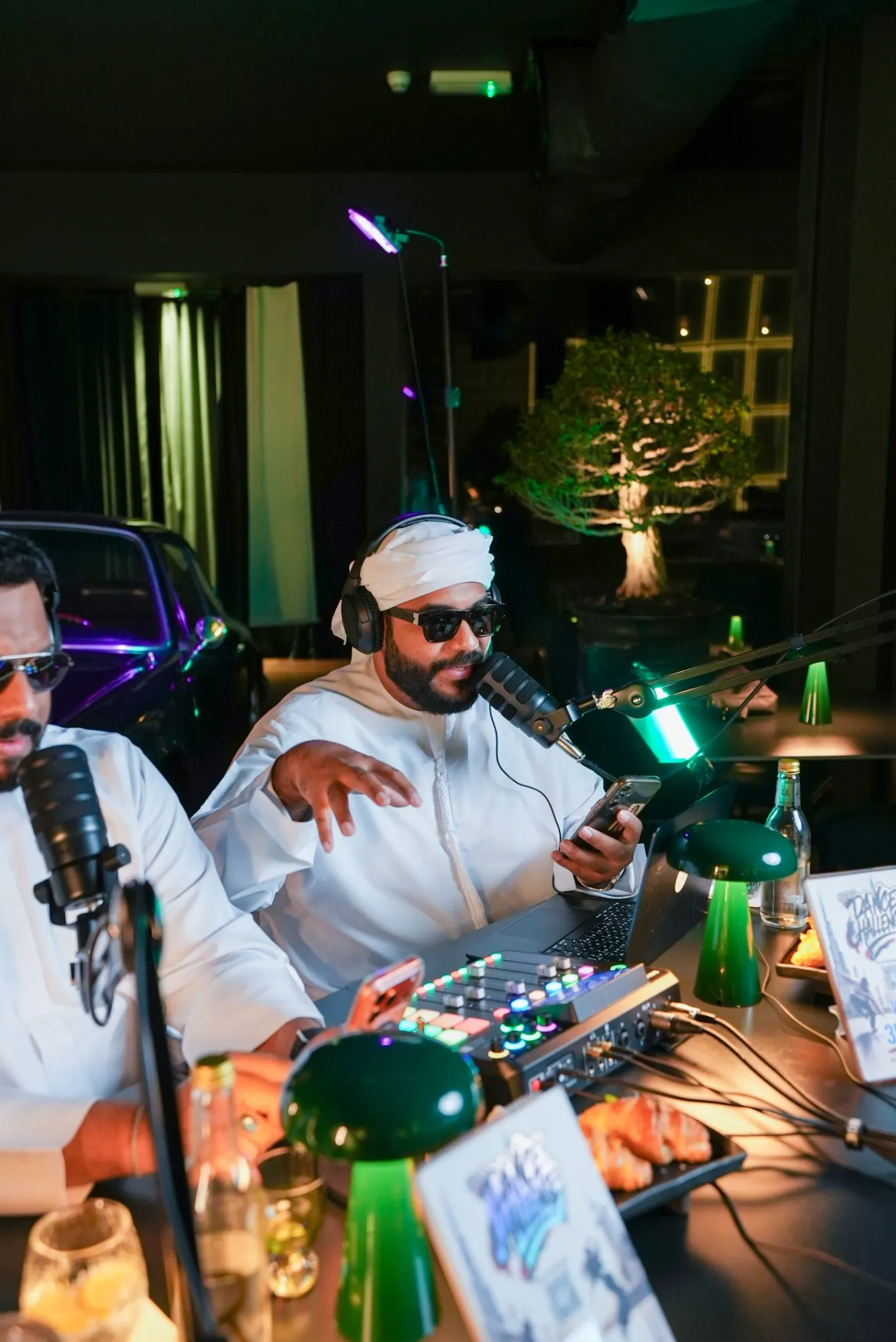 A podcast host in a white ghutra and sunglasses speaking into a microphone with a phone in hand, a bonsai and a purple-lit classic car behind