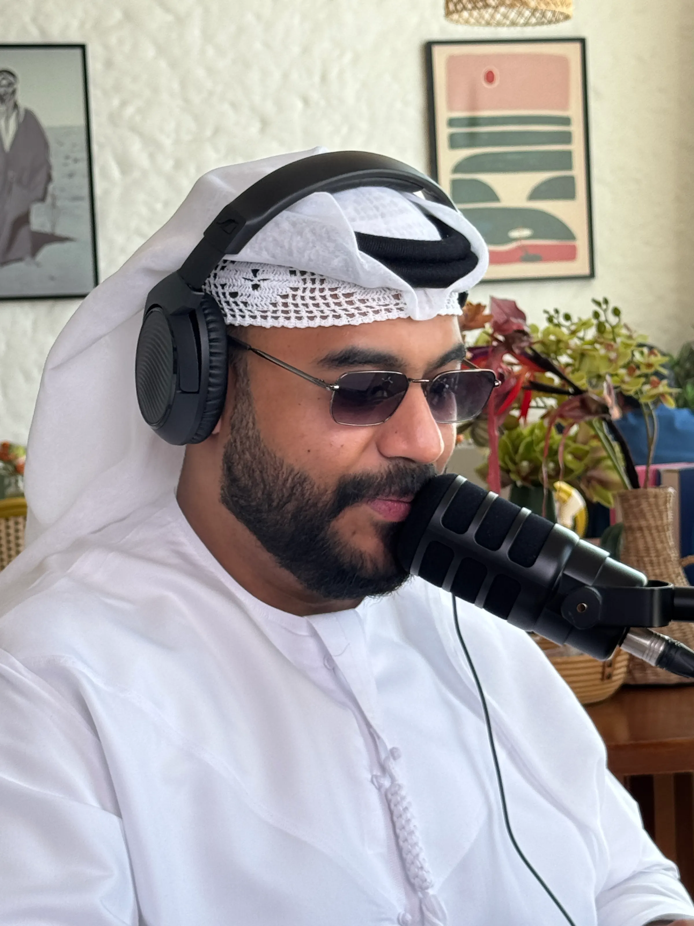 A close-up of a podcast host in a white ghutra, headphones and tinted glasses speaking into a microphone