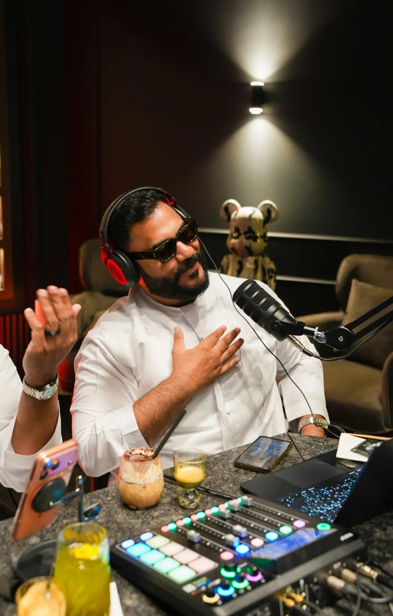 A podcast host in red-lit headphones and dark glasses with a hand on his heart at the microphone, a colourful mixer and drinks on the table