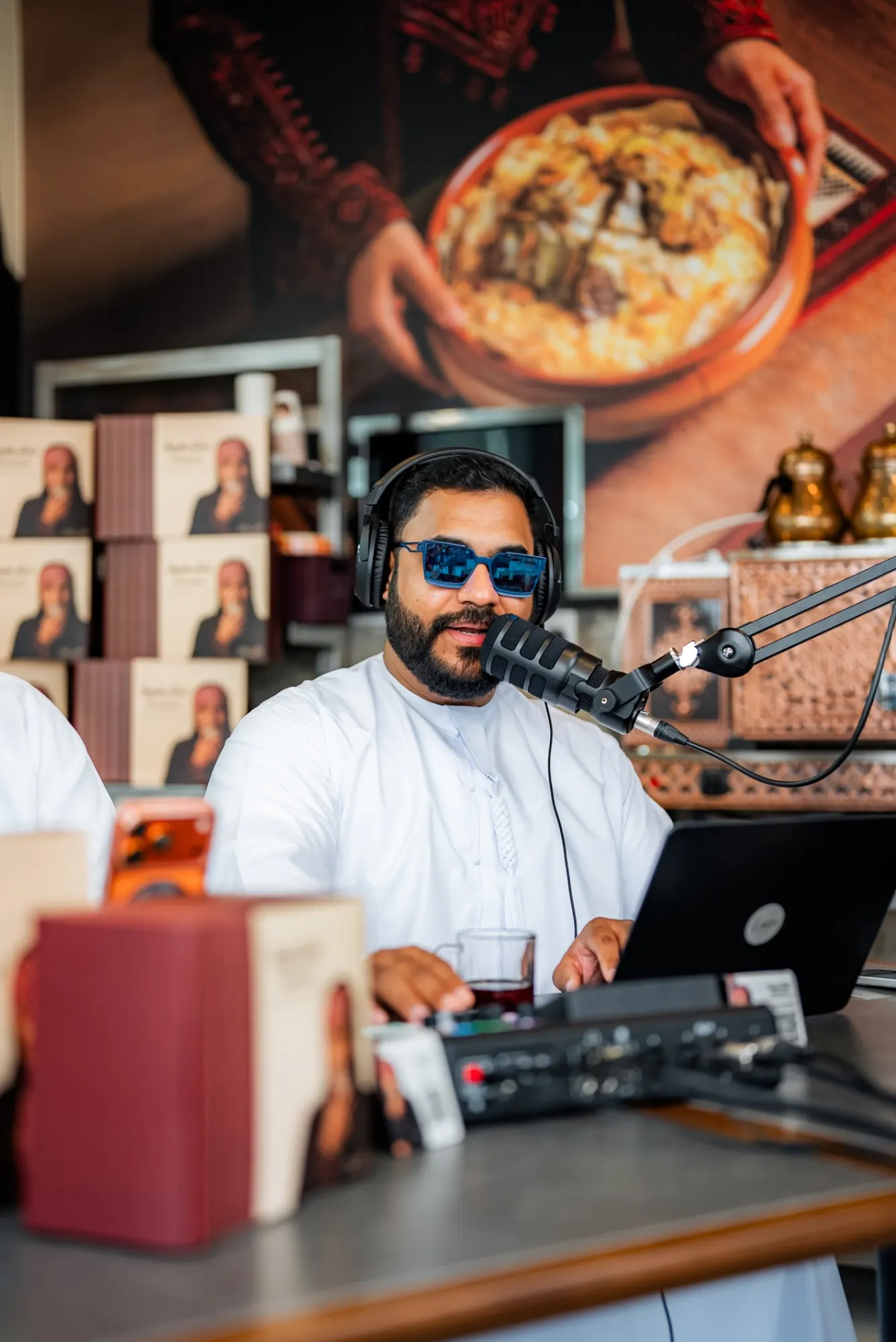 A podcast host in a white kandura and blue sunglasses speaking into a microphone at a restaurant counter, a large food mural behind him