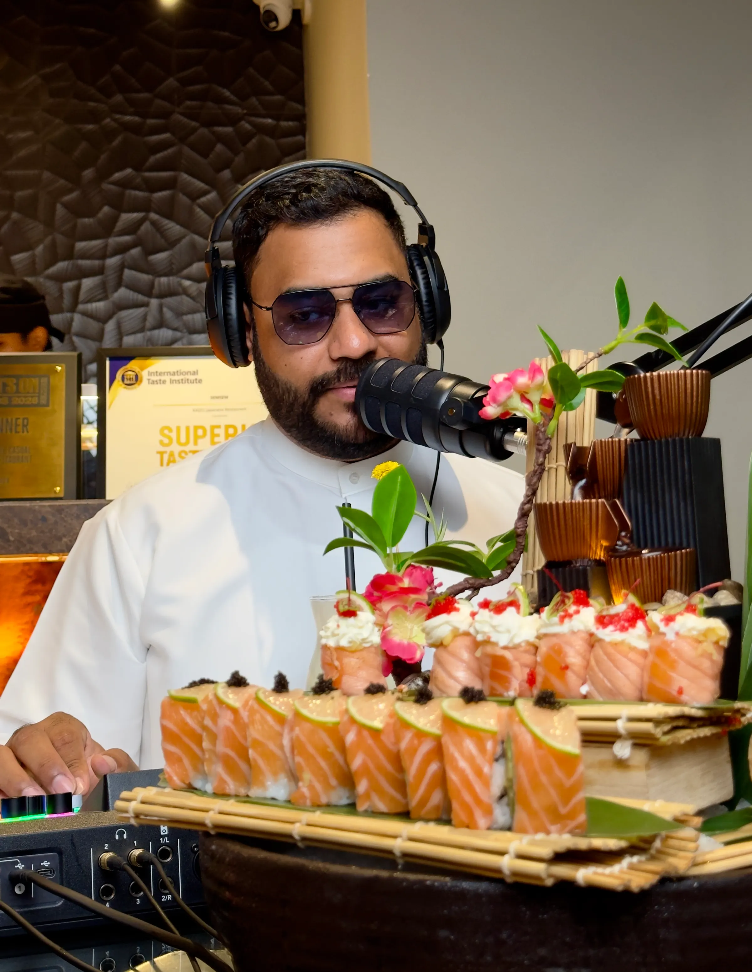 A podcast host in headphones and sunglasses behind a tall platter of salmon sushi rolls decorated with flowers