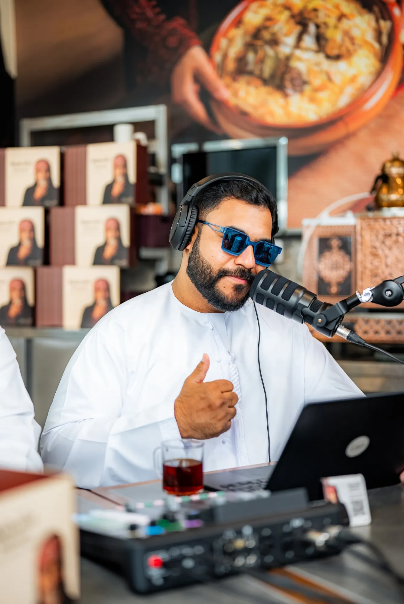 A podcast host in a white kandura, headphones and blue sunglasses giving a thumbs-up at a microphone, with a laptop, a mixer and a glass of tea in front of him