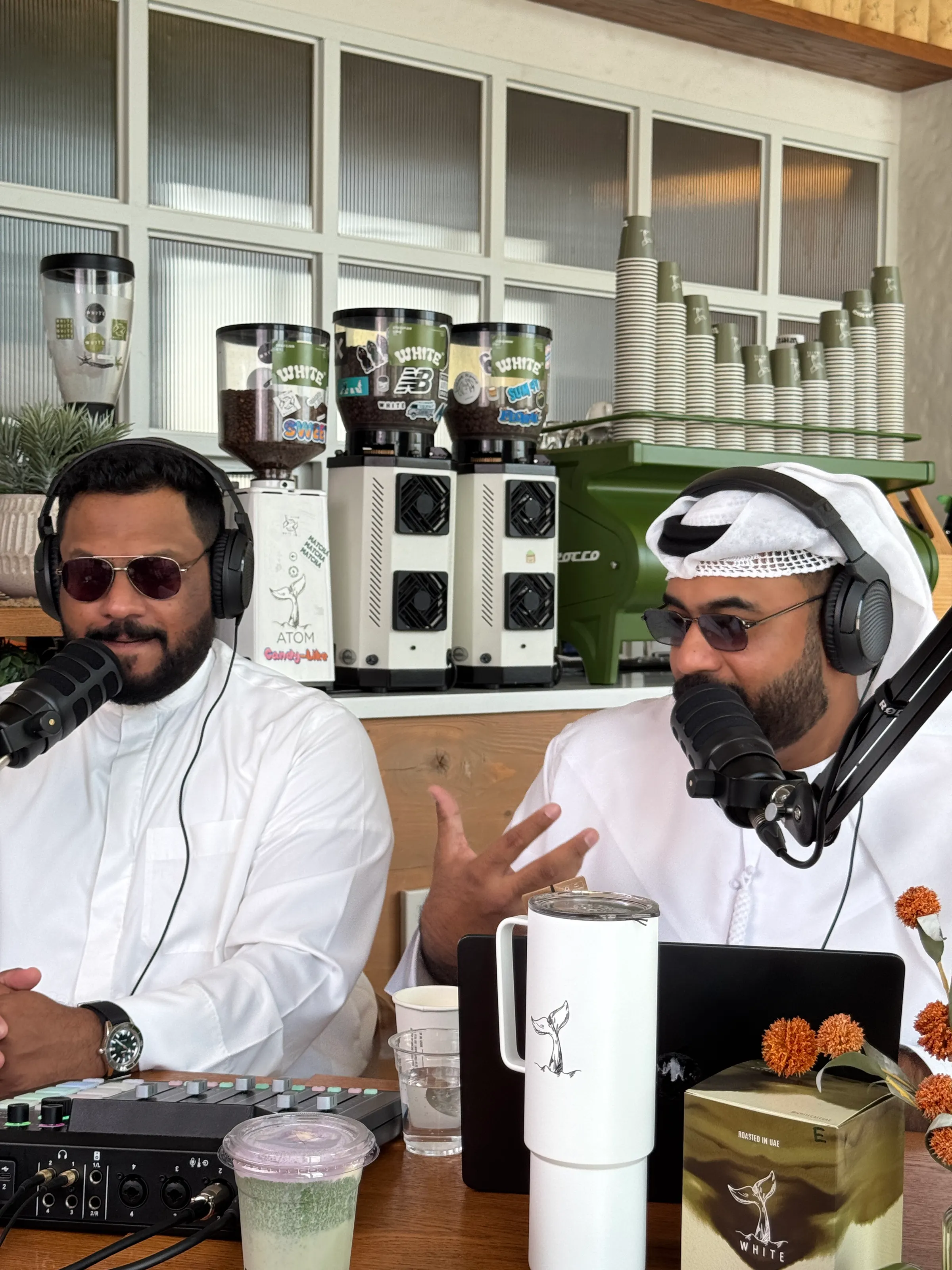 Two podcast hosts in white kanduras and headphones mid-conversation at a café counter, coffee grinders and stacked cups behind them