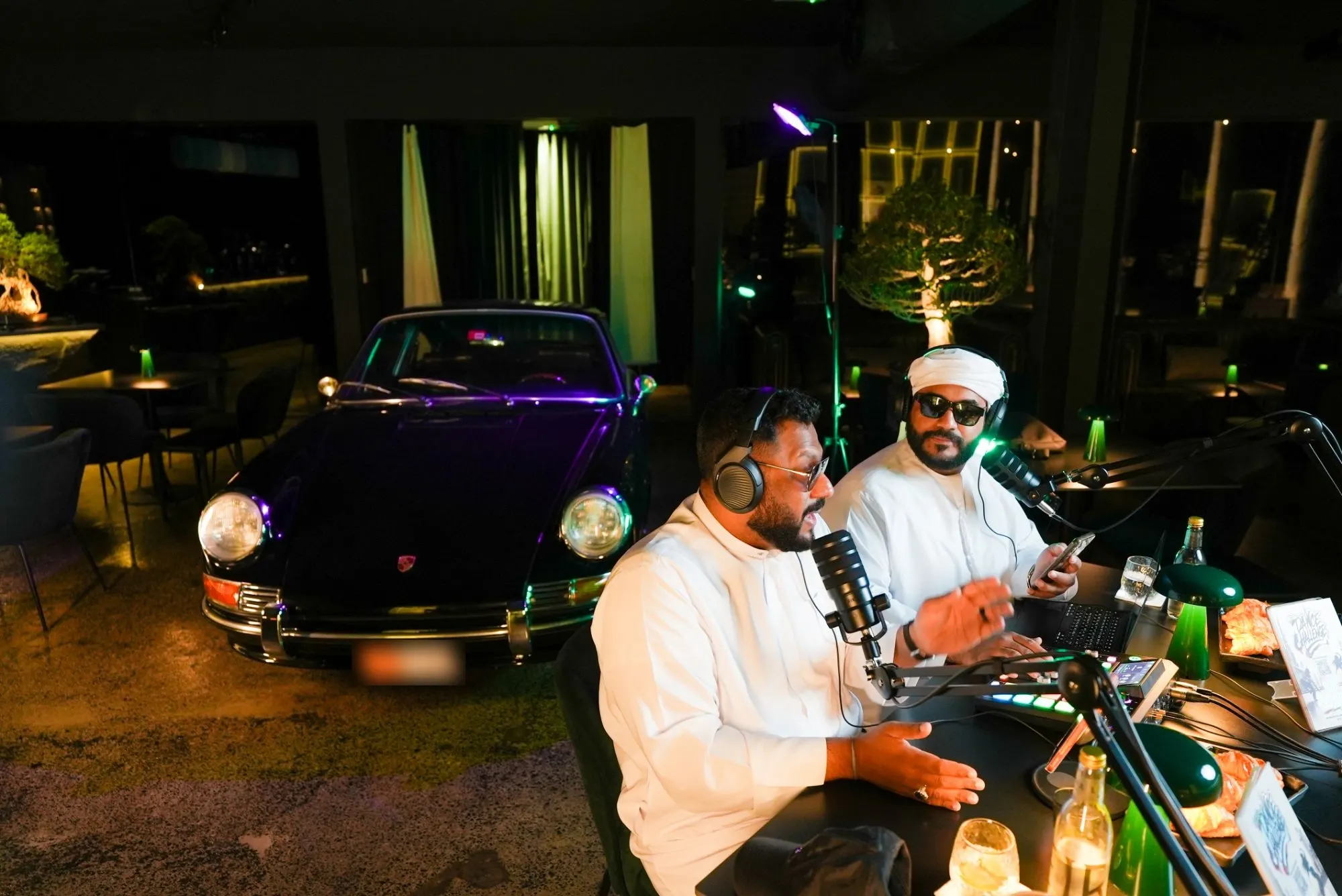 Two podcast hosts recording at a table beside a black classic sports car lit in purple and green, in a dark café