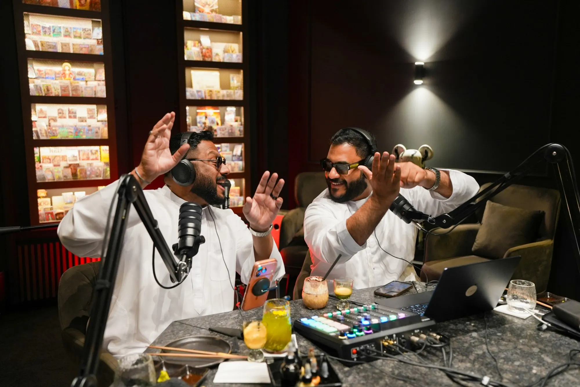 Two podcast hosts in white kanduras and headphones laughing and gesturing at their microphones in a dark lounge lined with lit display shelves