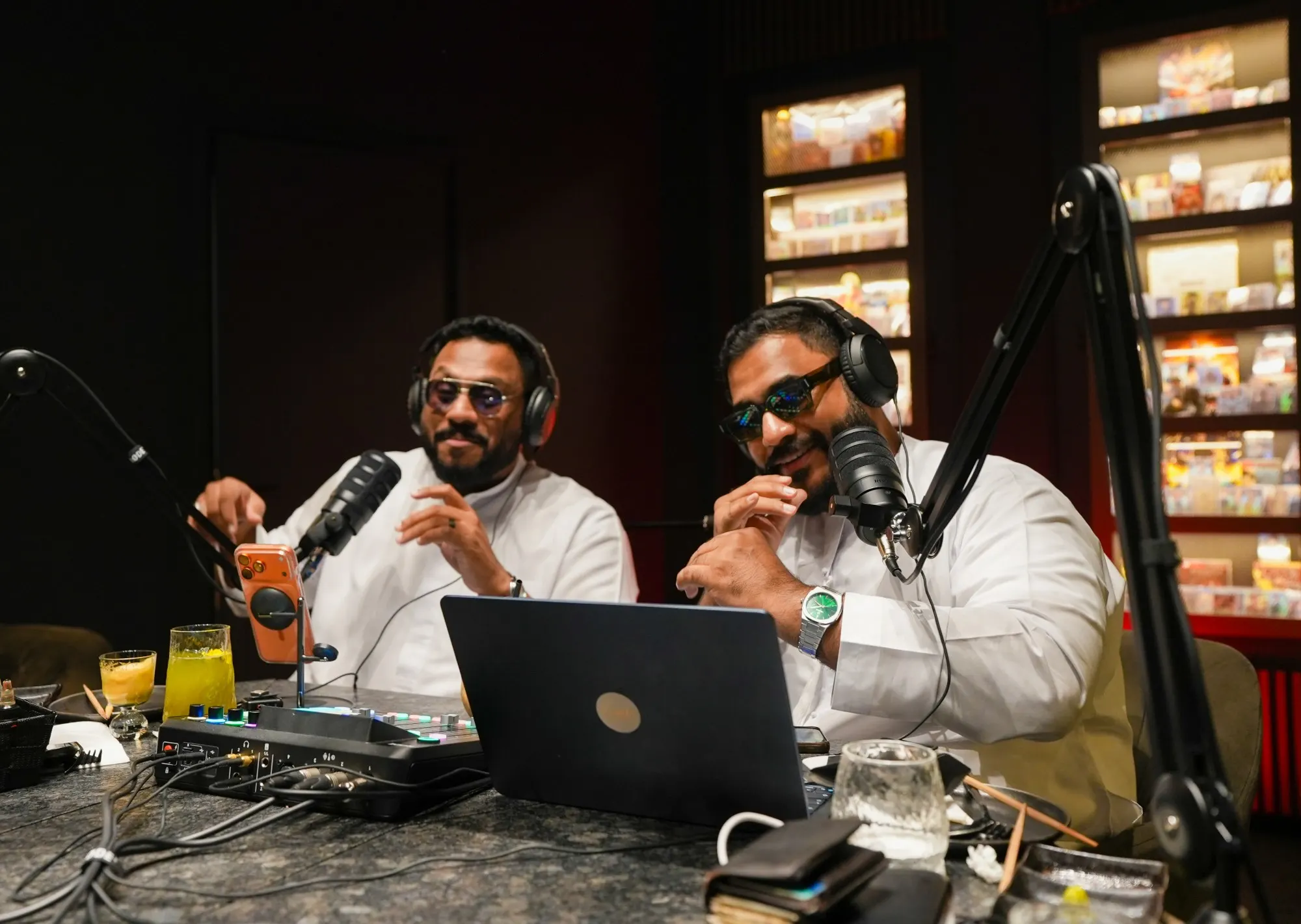 Two podcast hosts smiling at their microphones behind a laptop and a mixer, lit display shelves glowing behind them