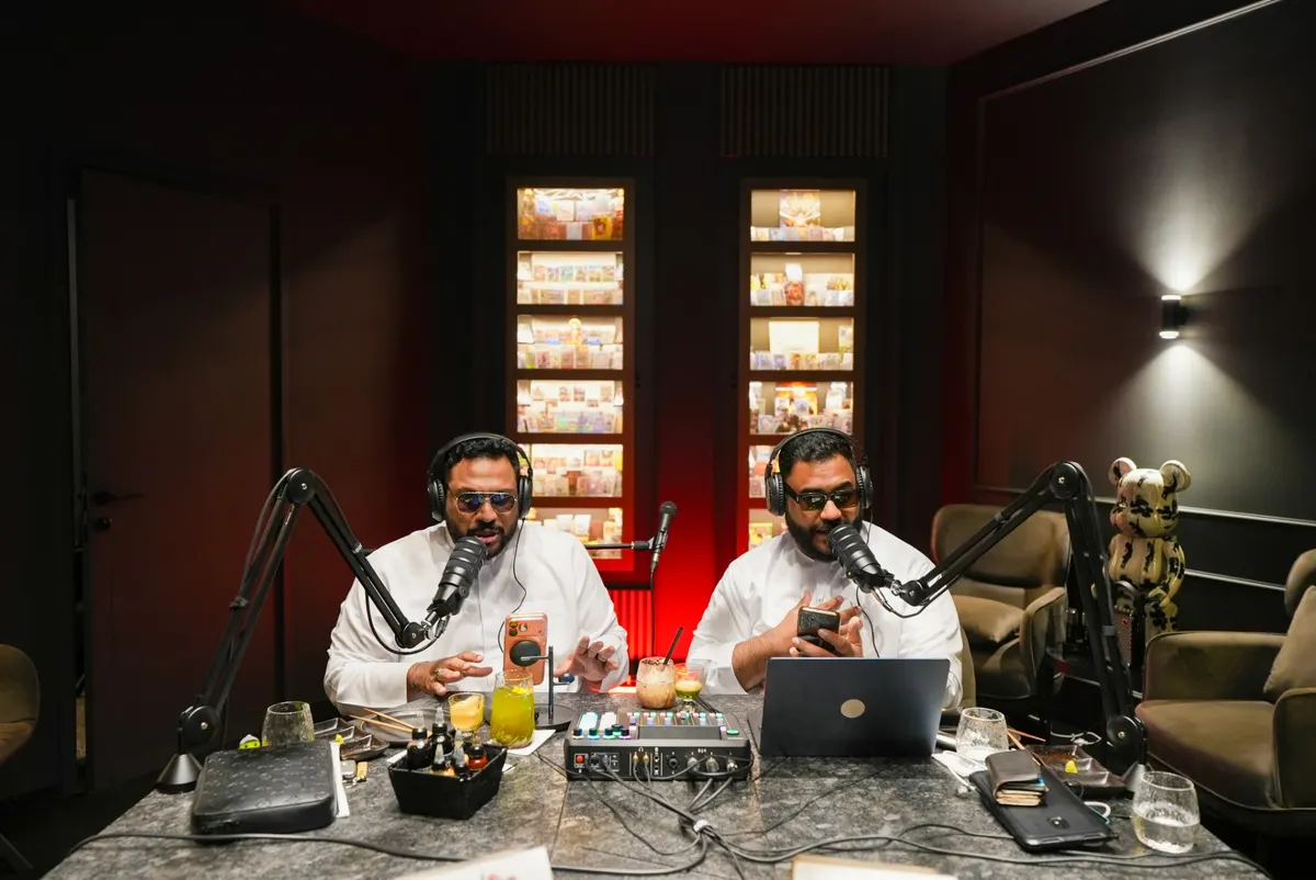 A symmetrical wide shot of two podcast hosts recording side by side, boom-arm microphones either side, red light glowing between two lit shelves