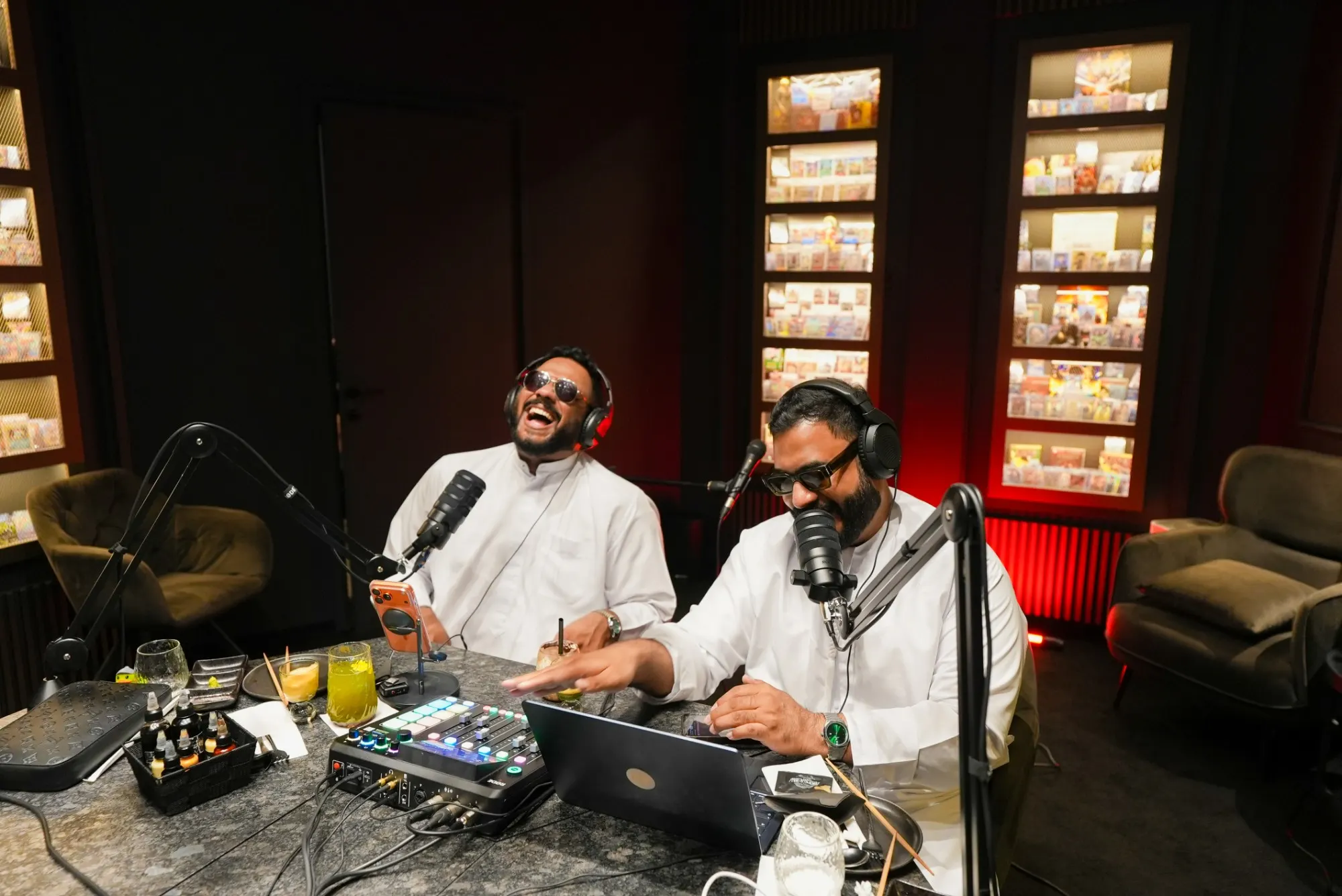 A wide view of two podcast hosts laughing at their microphones in a dark lounge with lit shelves, armchairs and red accent light