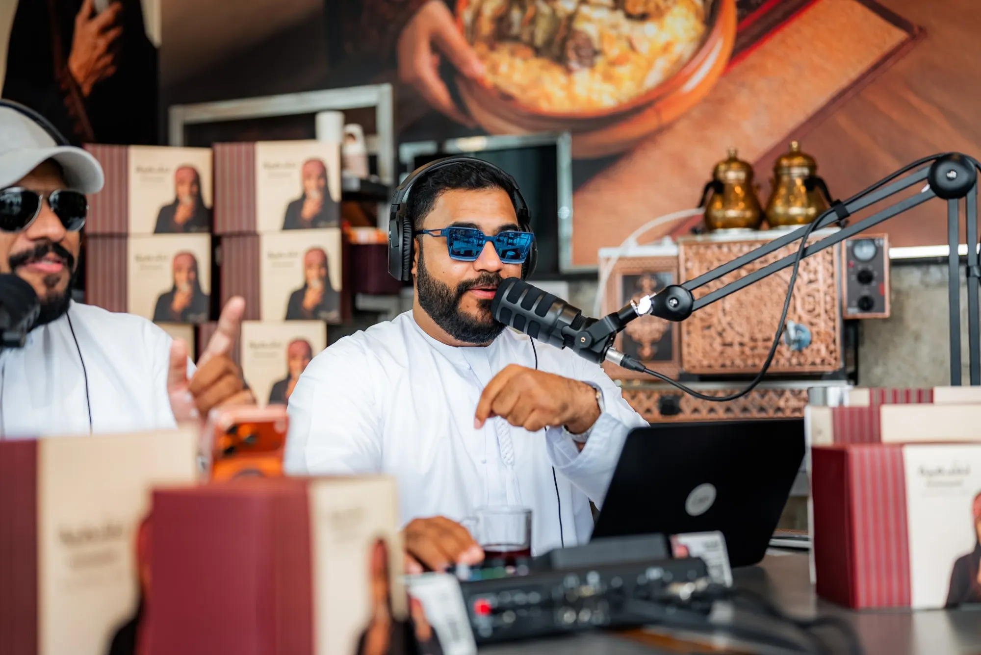 Two podcast hosts in white kanduras recording at a restaurant counter, one speaking into a boom-arm microphone behind a laptop and a mixer