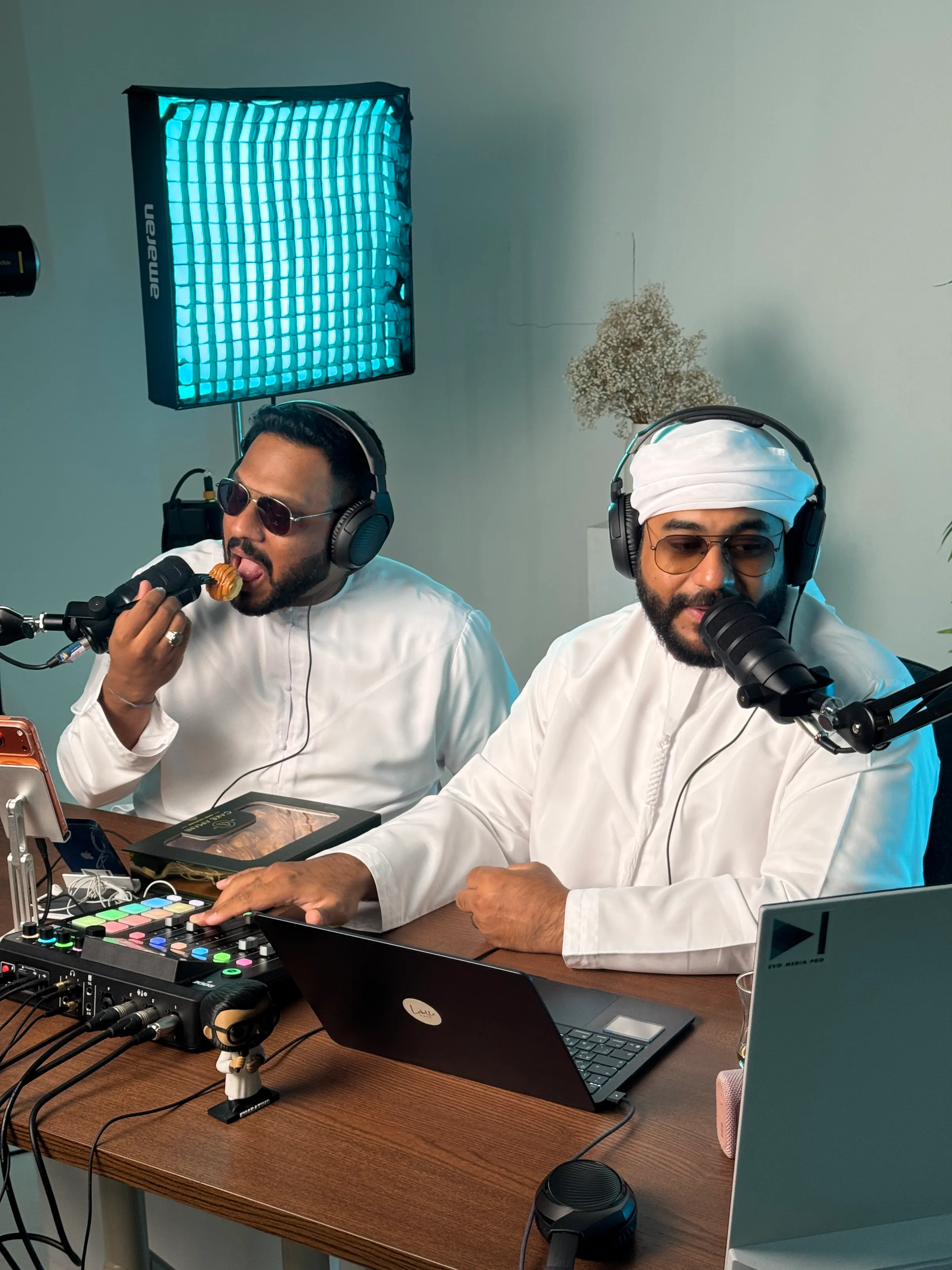 Two podcast hosts at a wooden desk under a teal softbox, one tasting a pastry at the mic while the other works the mixer