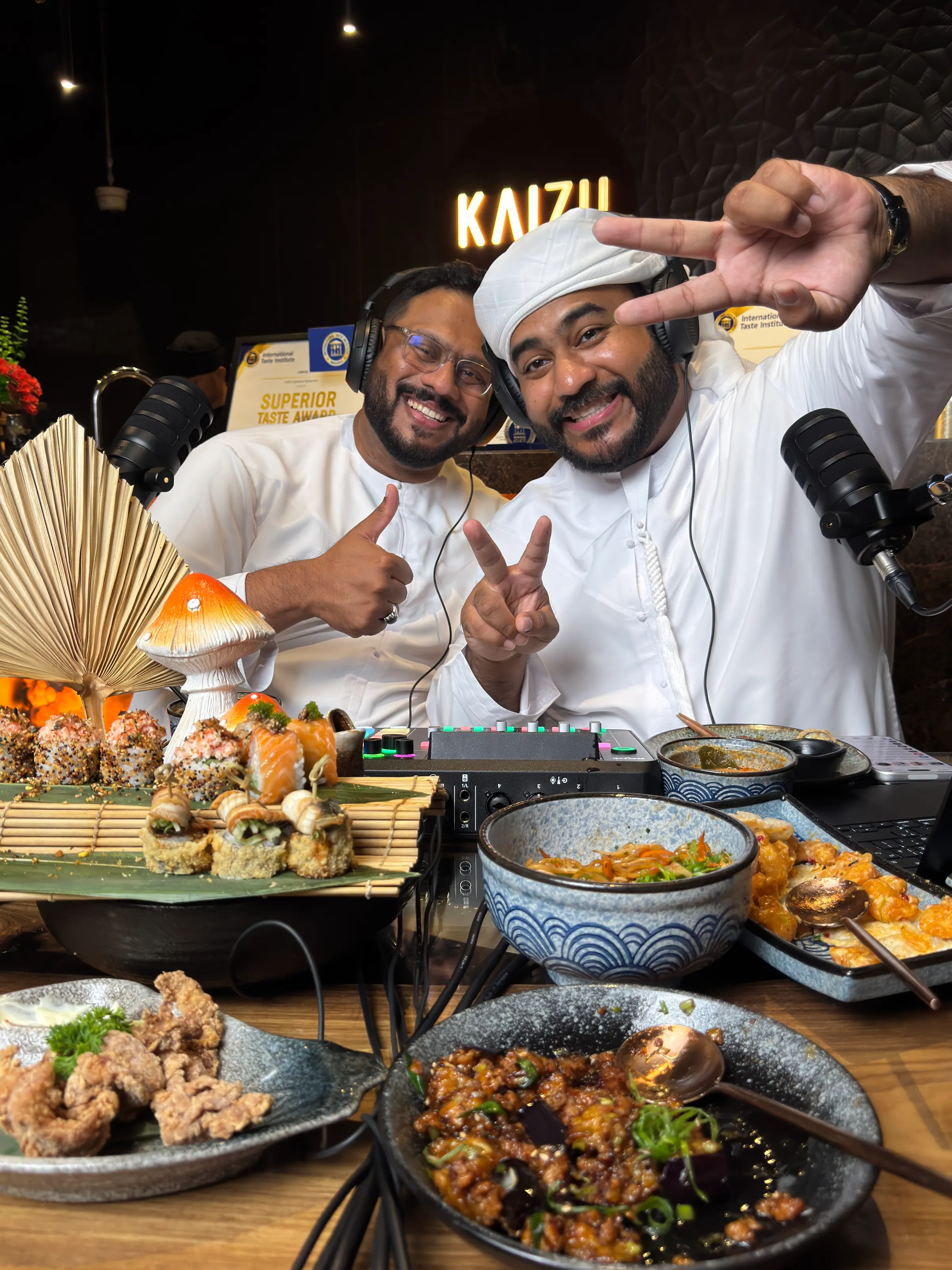 Two podcast hosts in white kanduras posing with a thumbs-up and a peace sign behind a spread of sushi and small plates