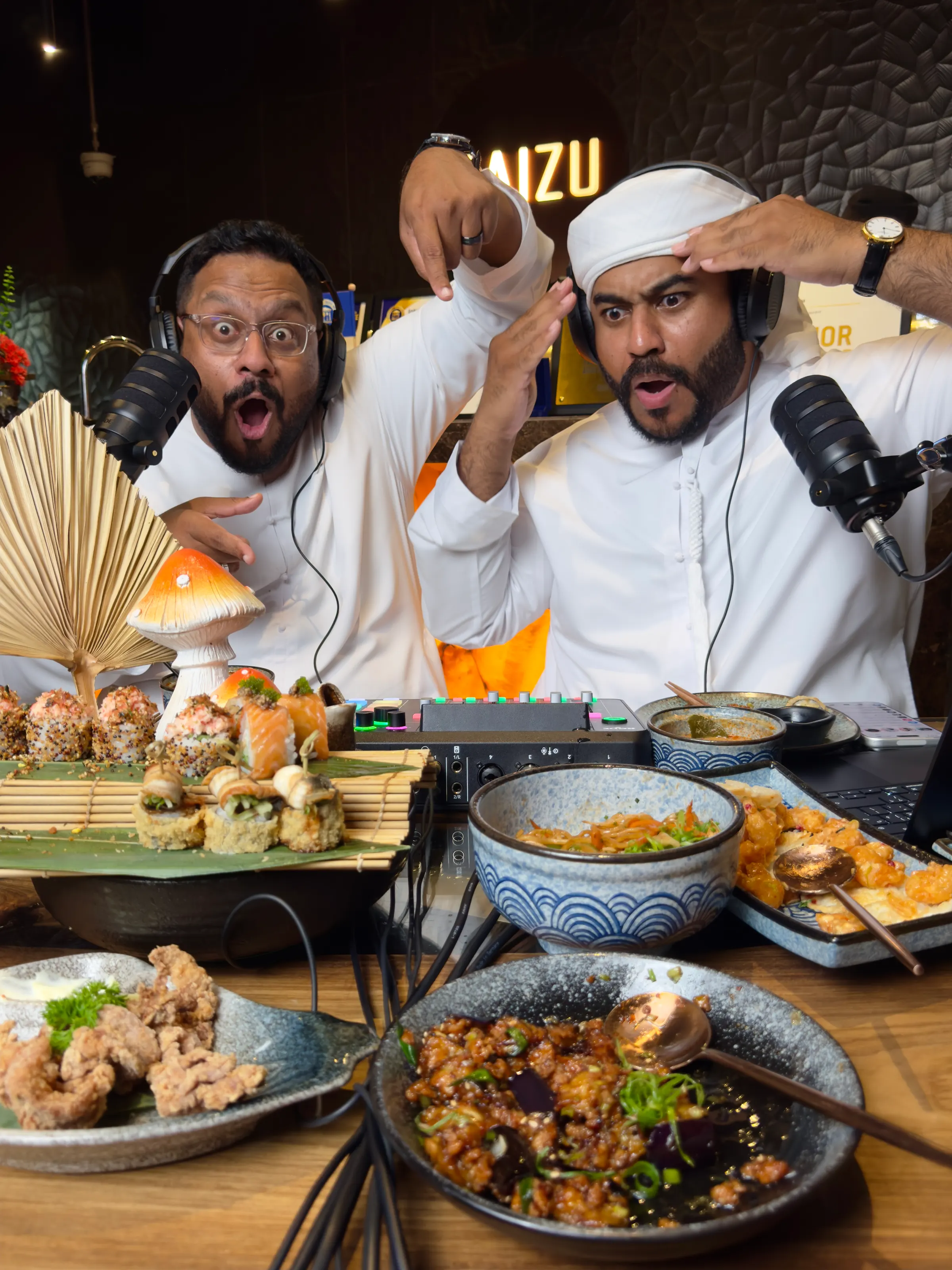 Two podcast hosts pulling surprised faces at their microphones behind a spread of sushi and small plates