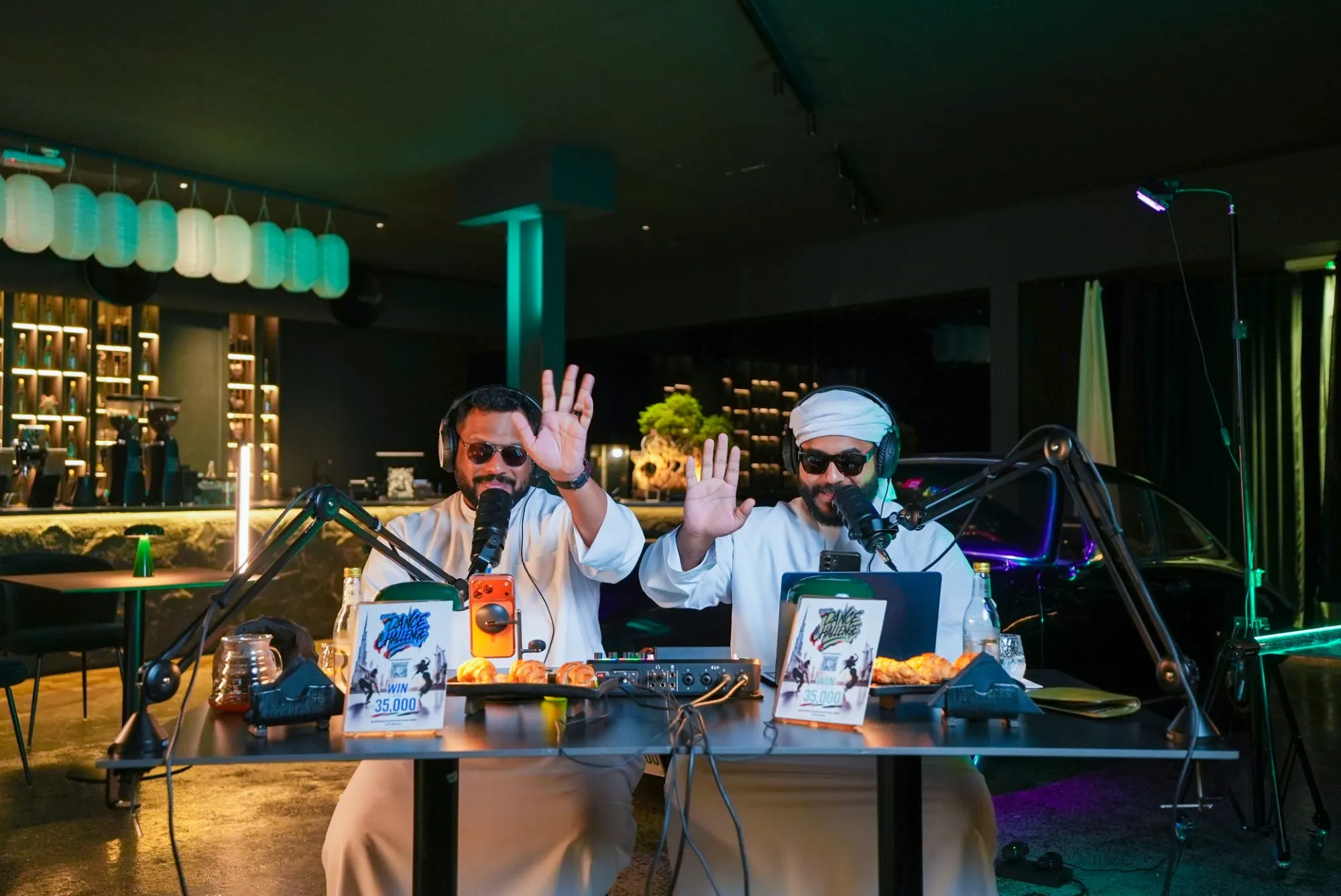 Two podcast hosts waving to camera at a table set up in a dark café, paper lanterns glowing over the bar behind them