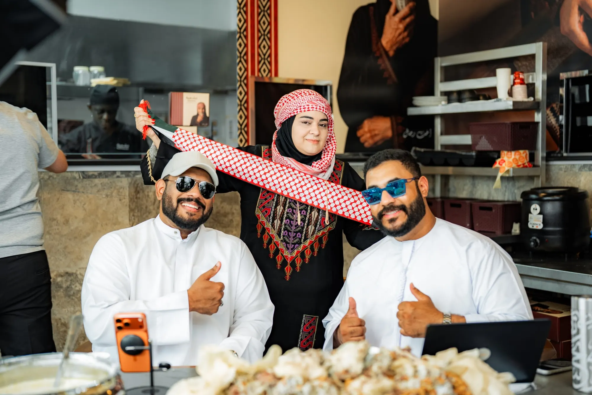 The two podcast hosts giving thumbs-ups at their table while a guest in an embroidered dress and keffiyeh holds a keffiyeh-print scarf above them