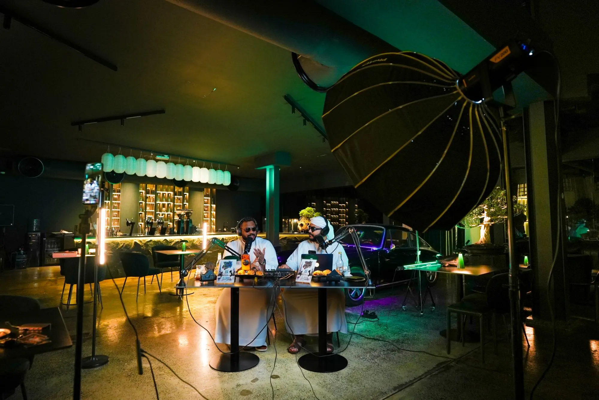 Behind the scenes of a podcast recording in a dark café: a large parabolic softbox, a phone on a rig and light tubes framing the two hosts at their table
